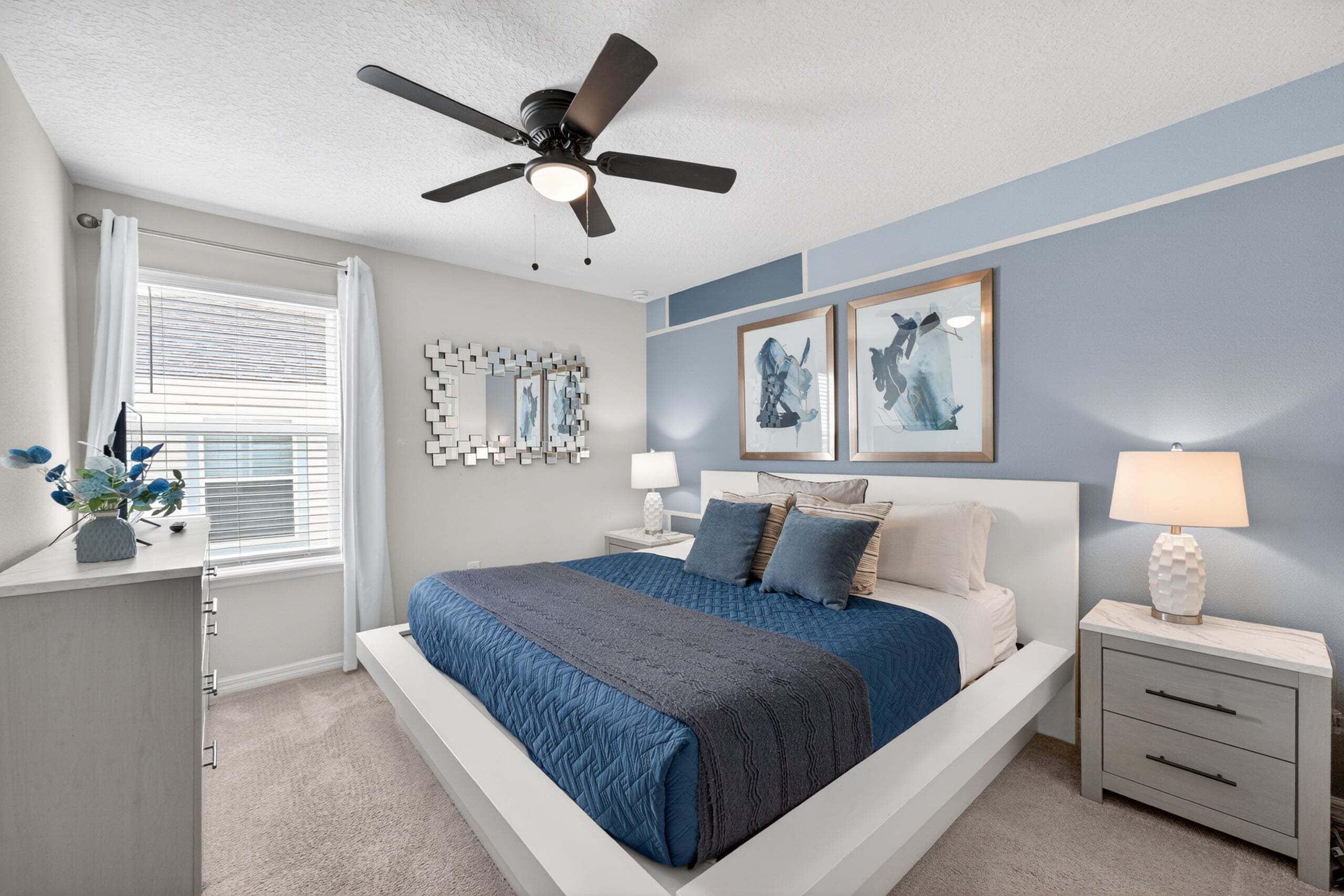 A bedroom with a bed, bedside tables, a ceiling fan, a window with blinds, and wall art.