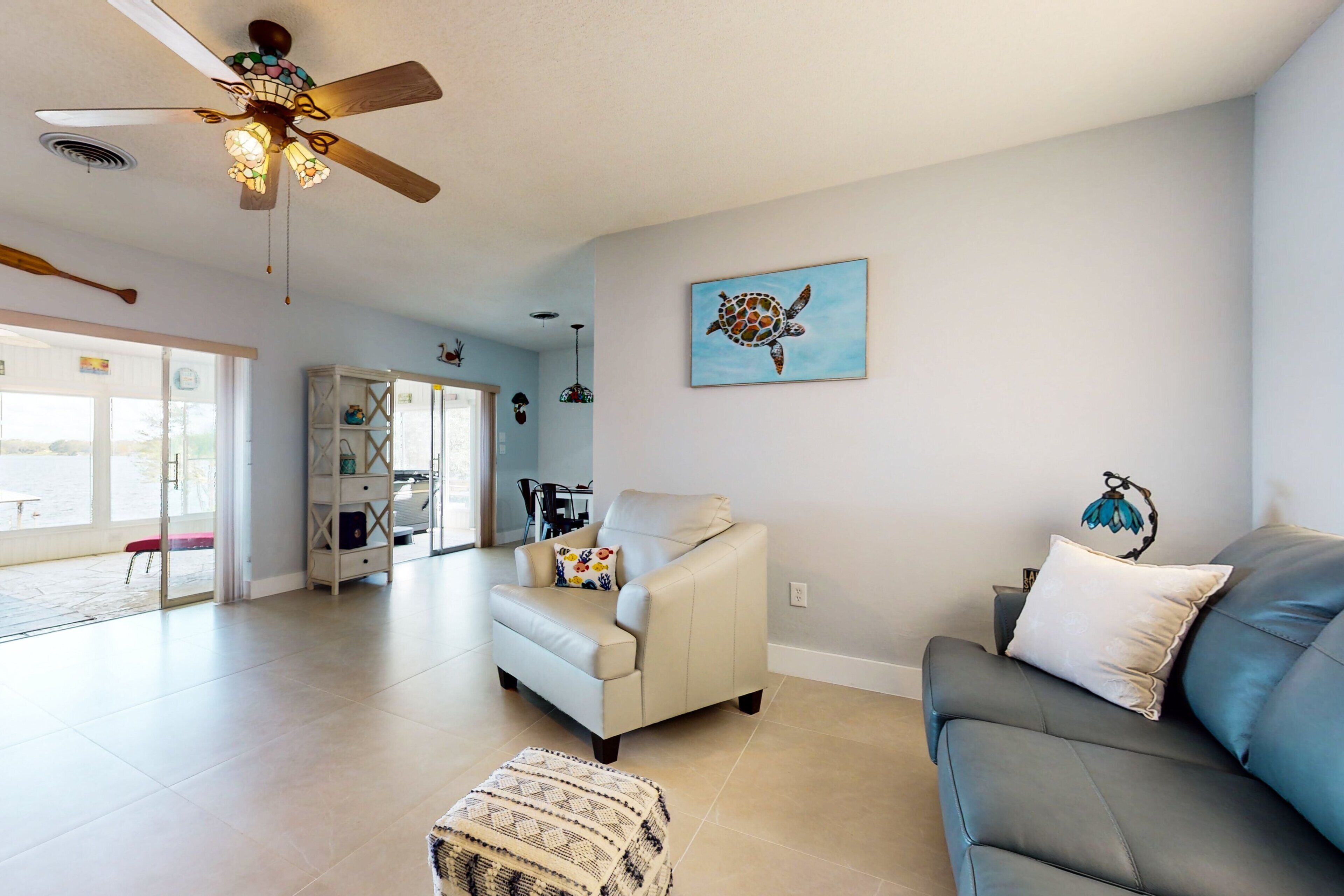 A spacious living room with a ceiling fan, a beige armchair, a gray sofa, and a wall-mounted turtle painting.