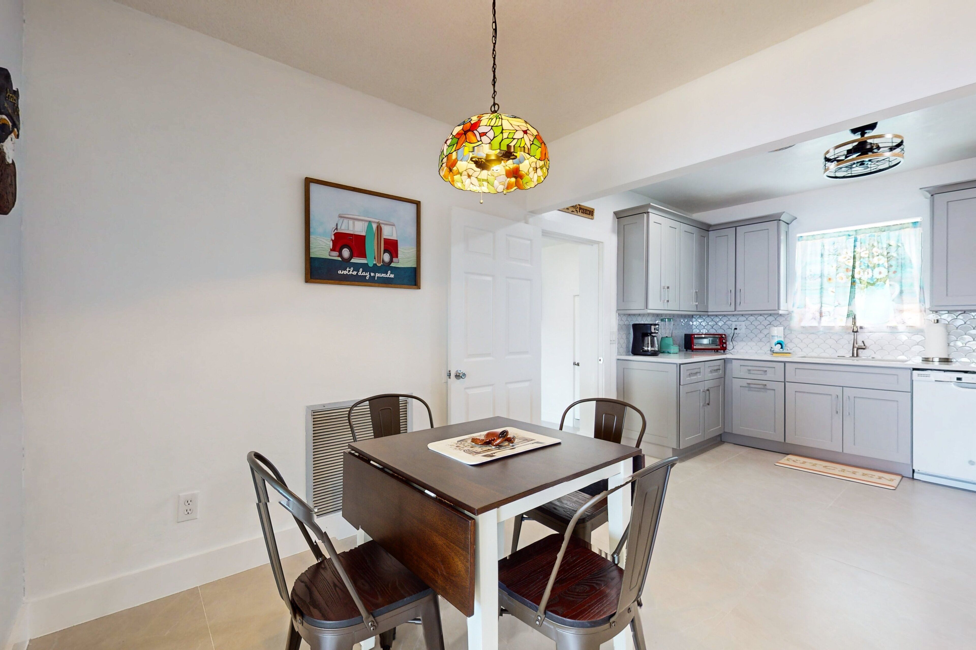 A dining area with a table and chairs, a hanging light fixture, a kitchen with cabinets and appliances, and a door leading to another room.