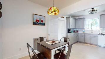 A dining area with a table and chairs, a hanging light fixture, a kitchen with cabinets and appliances, and a door leading to another room.