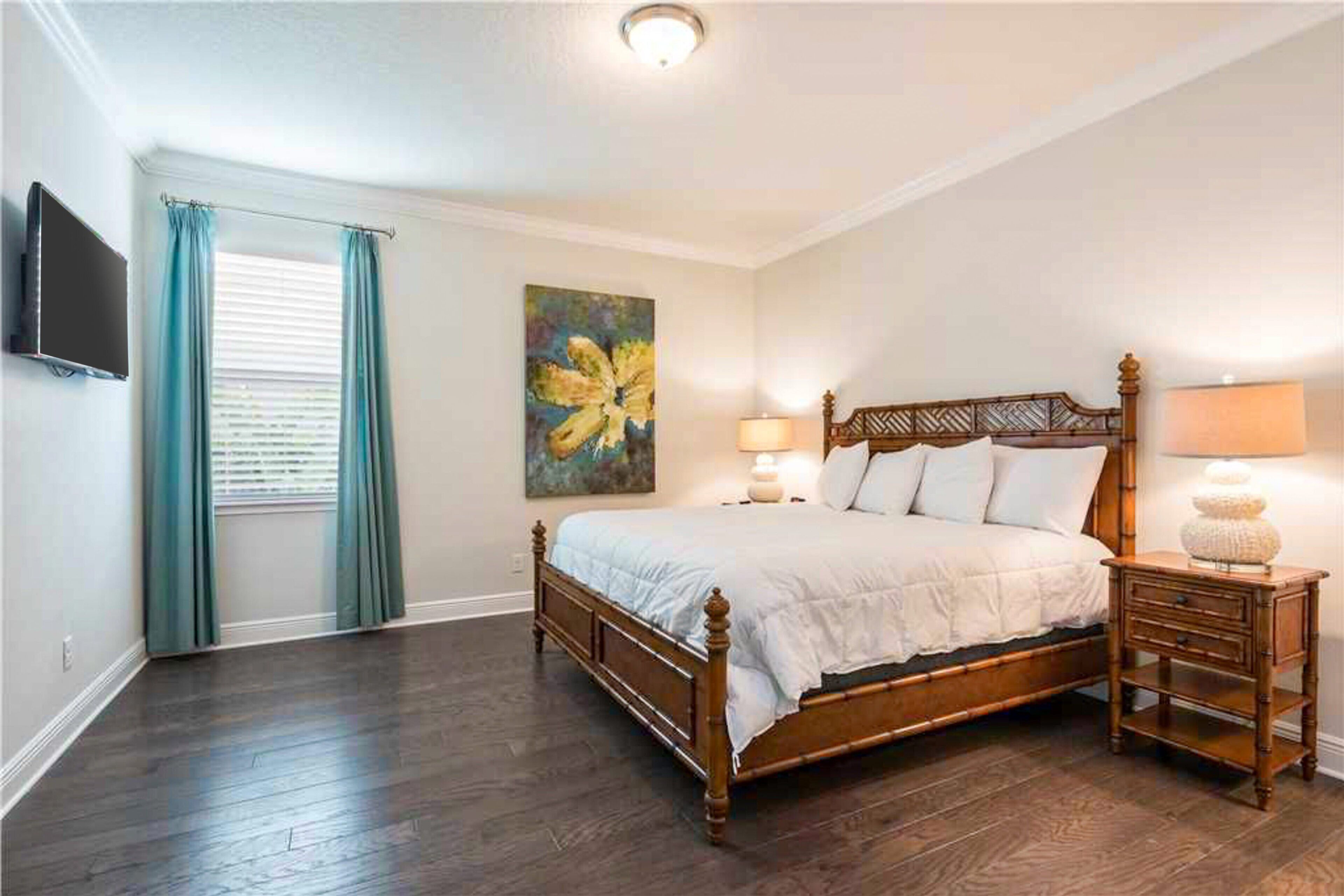 A bedroom with a wooden bed, a nightstand, a lamp, a painting, and a TV on the wall.