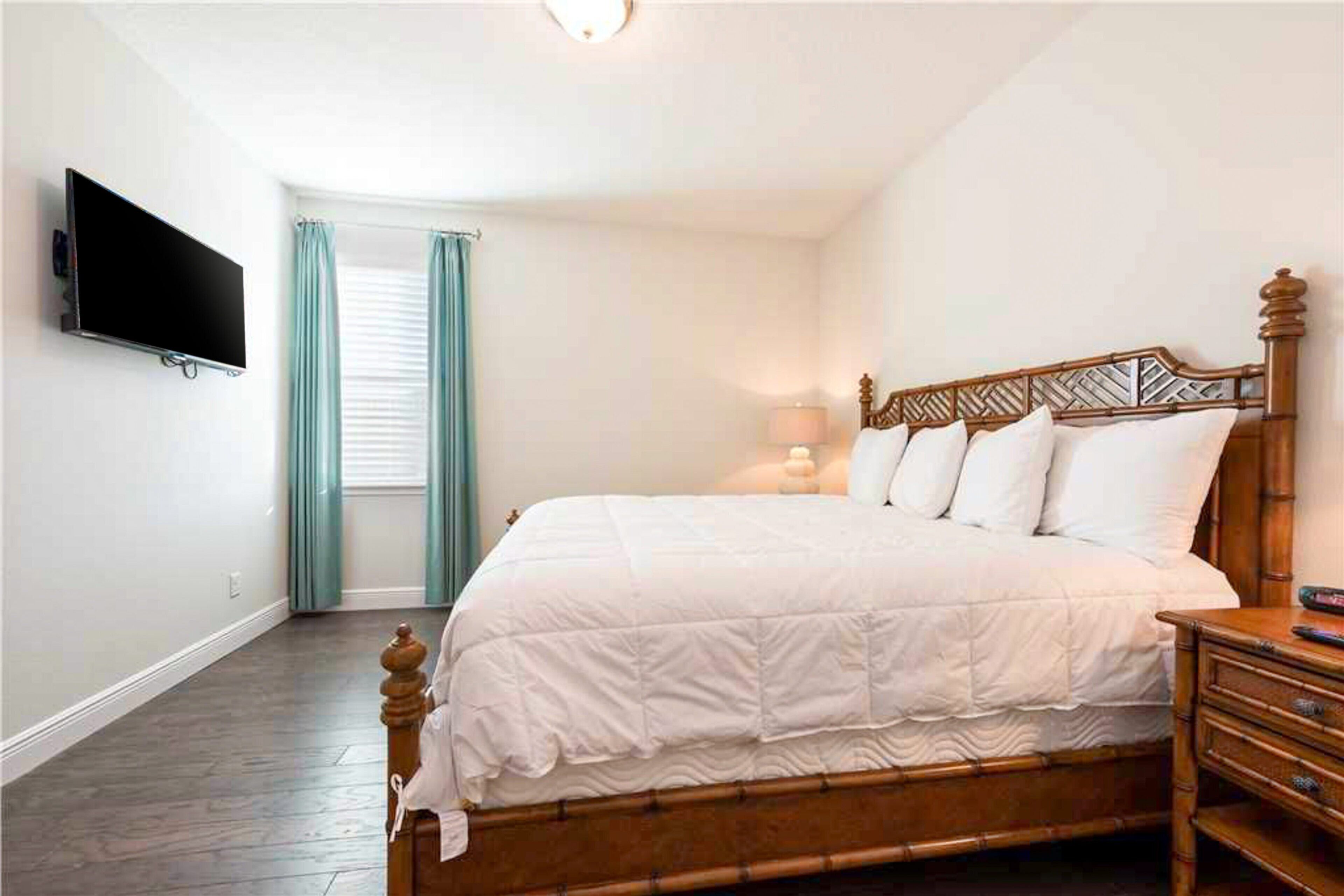 A bedroom with a wooden bed, a television mounted on the wall, a window with curtains, and a nightstand with a lamp.
