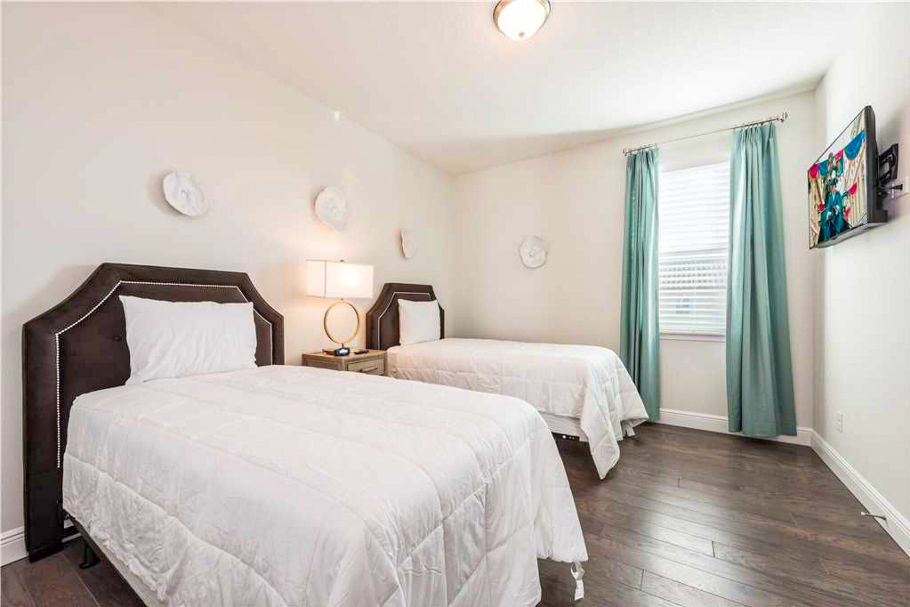 Une chambre d’hôtel avec deux lits, une table de chevet avec une lampe, une fenêtre avec des rideaux et un téléviseur fixé au mur.
