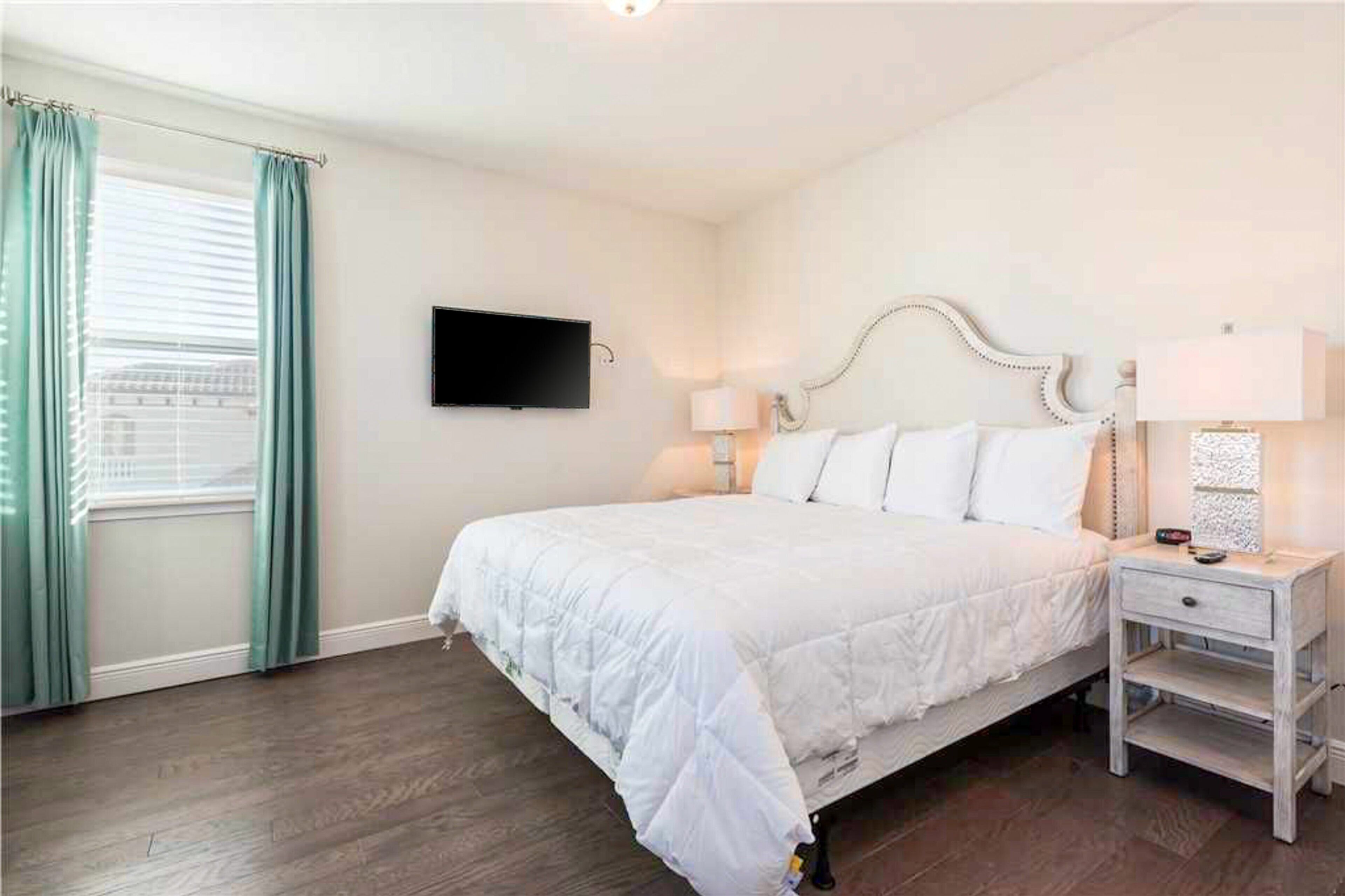 Un dormitorio con una cama, un televisor montado en la pared, una mesita de noche con una lámpara y una ventana con cortinas.