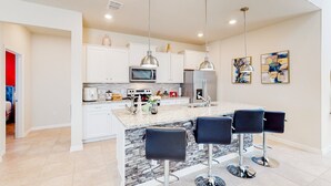 Una cucina moderna con mobili bianchi, un'isola in granito con sgabelli da bar, elettrodomestici in acciaio inossidabile e illuminazione a sospensione.