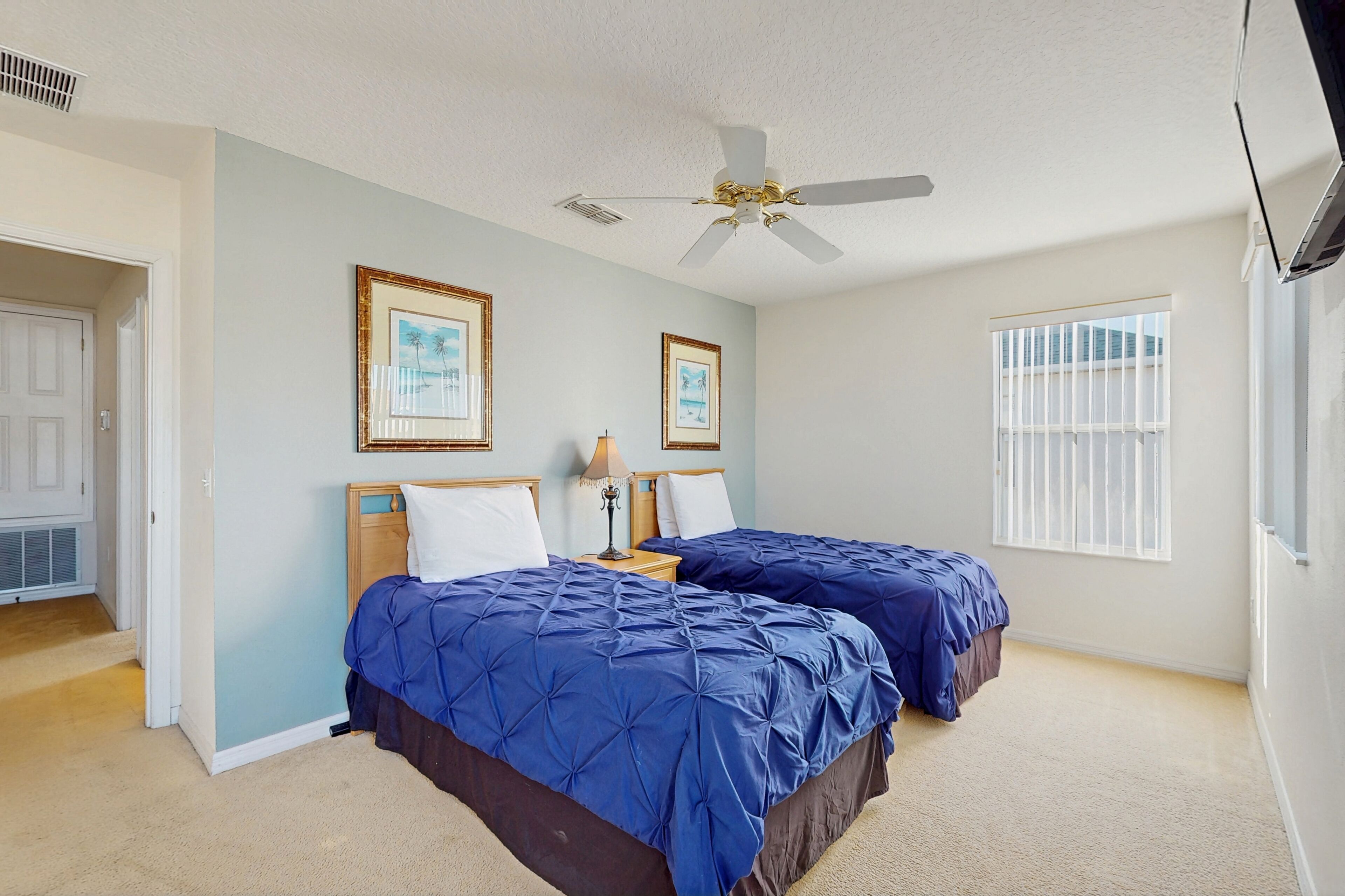 A bedroom with two beds, a ceiling fan, framed pictures on the wall, and a window with blinds.