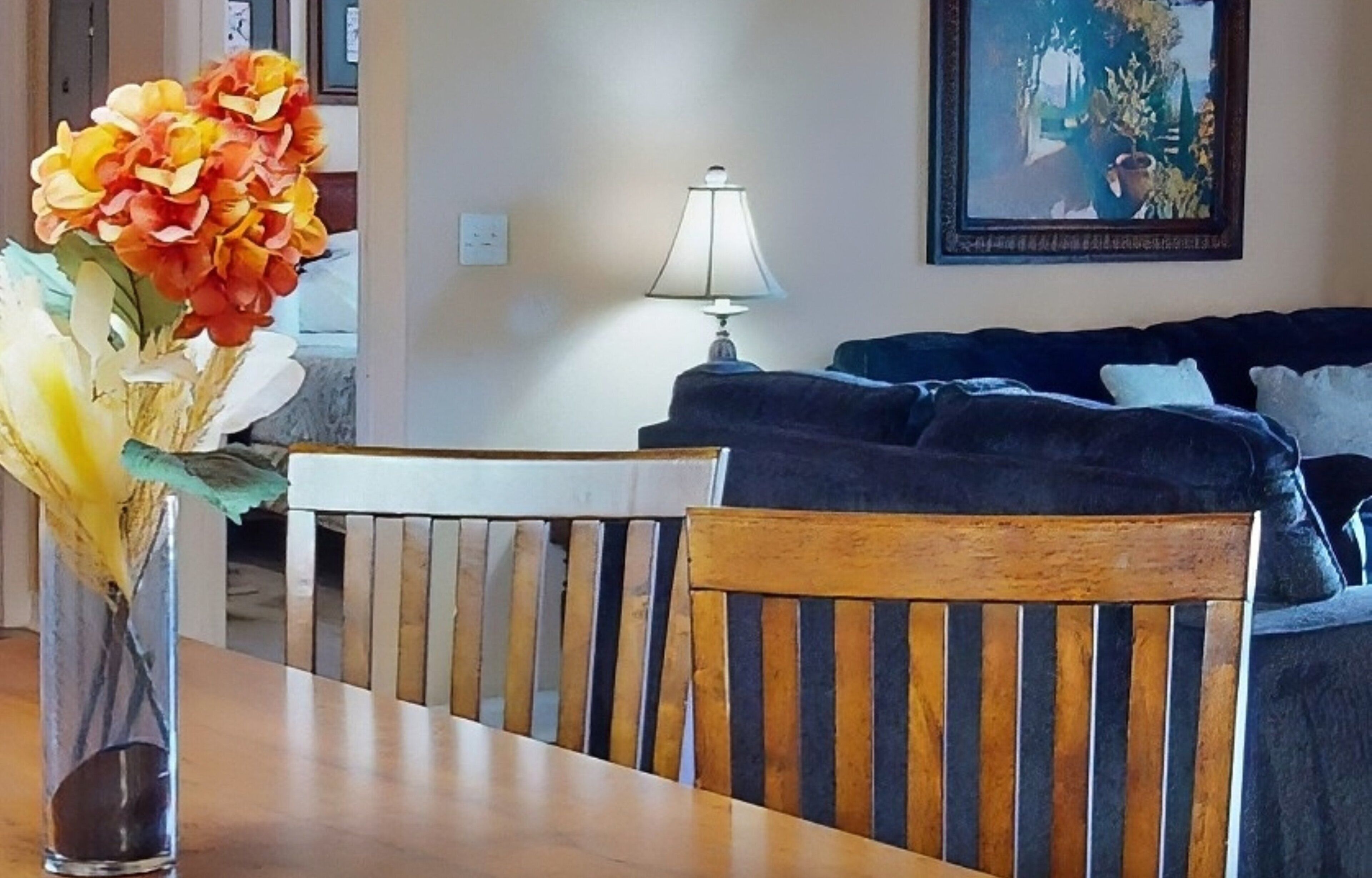 A dining area with a wooden table and chairs, a vase with flowers, a lamp, and a couch in the background.