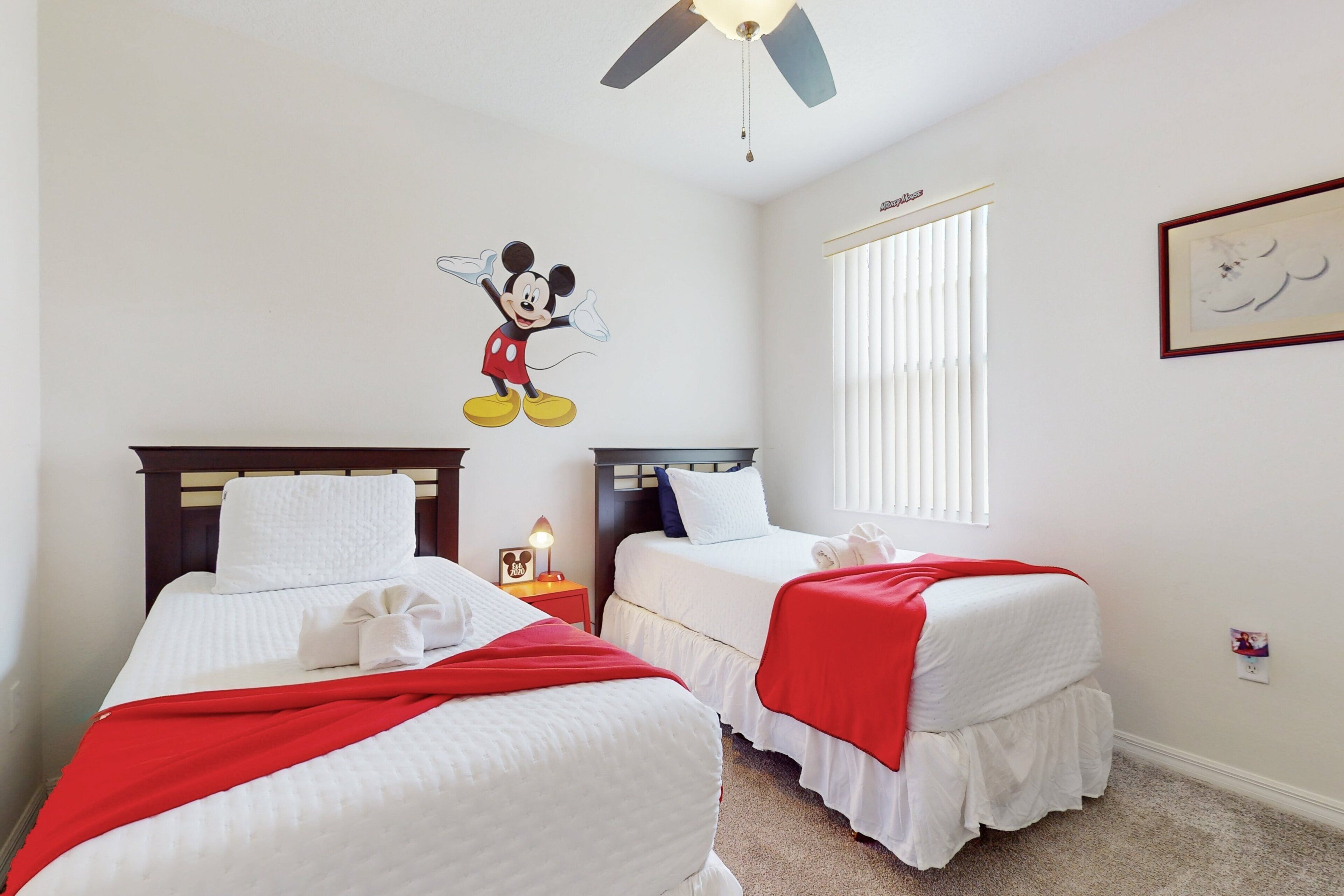 A hotel room with two beds, a ceiling fan, a window with blinds, and a Mickey Mouse wall decal.