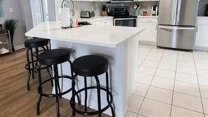 A modern kitchen with white cabinets, a large island with stools, stainless steel appliances, and a tiled floor. - Single-story Home With Private Pool, Wifi, Central AC, WD and Full Kitchen (Haines City)