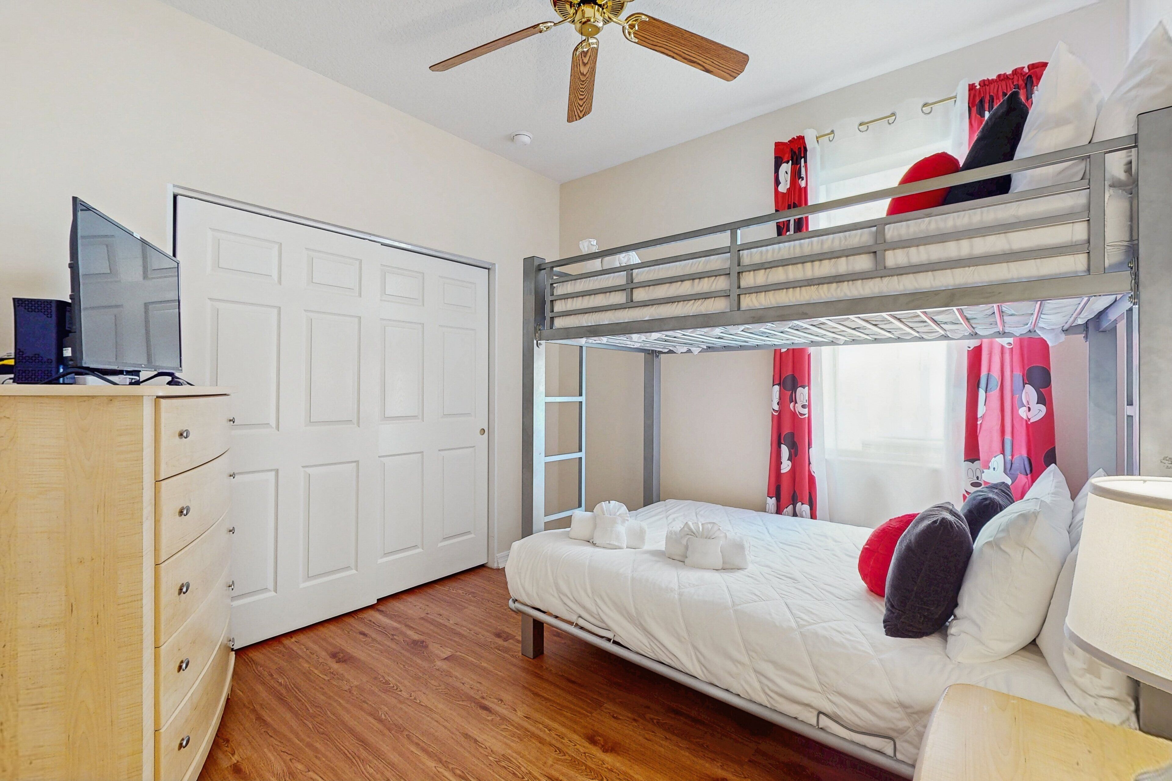 A bunk bed room with a wooden dresser, a ceiling fan, and a window with curtains.