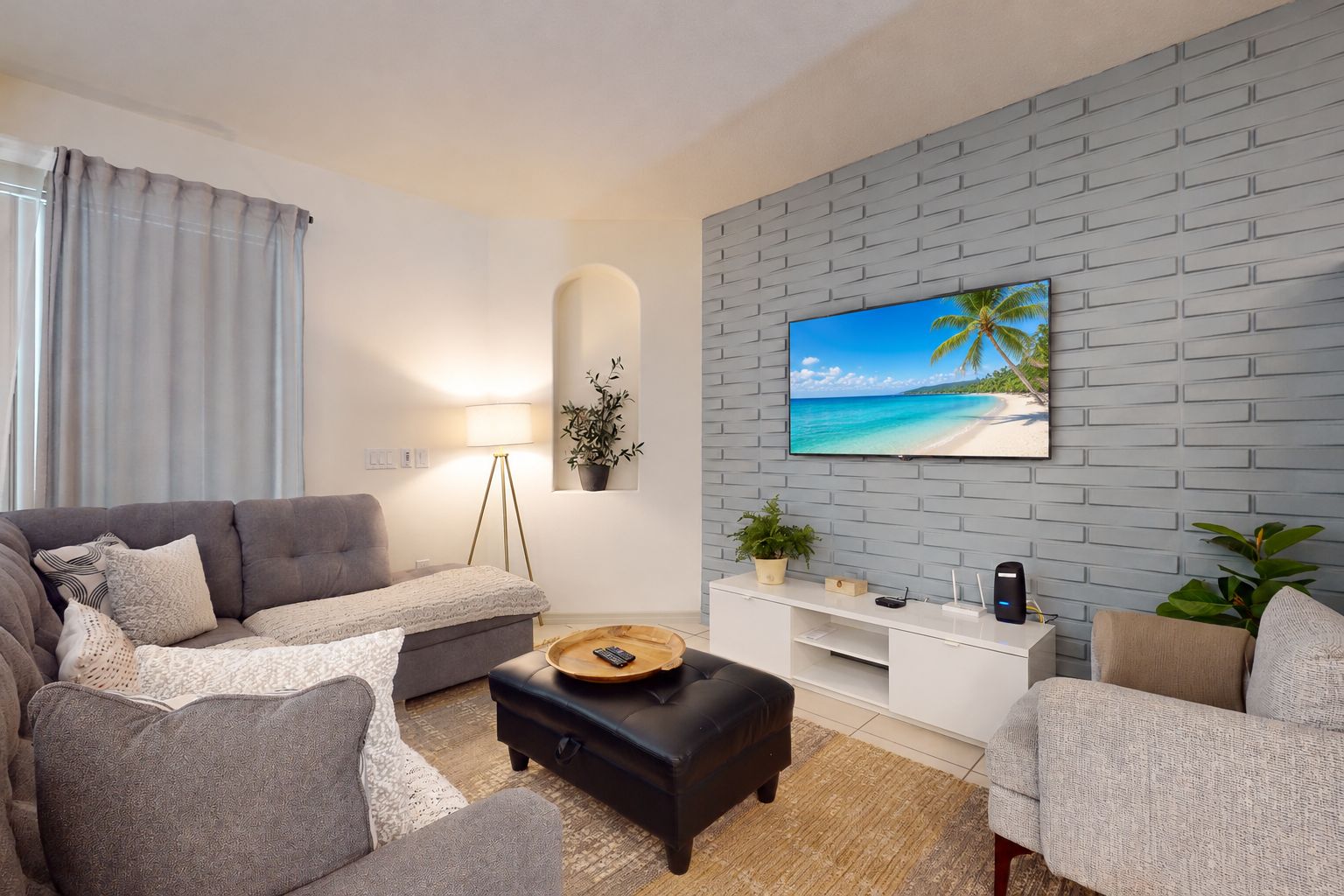 A modern living room with a grey sofa, a black ottoman, a white entertainment unit, and a large TV mounted on a brick-patterned wall.