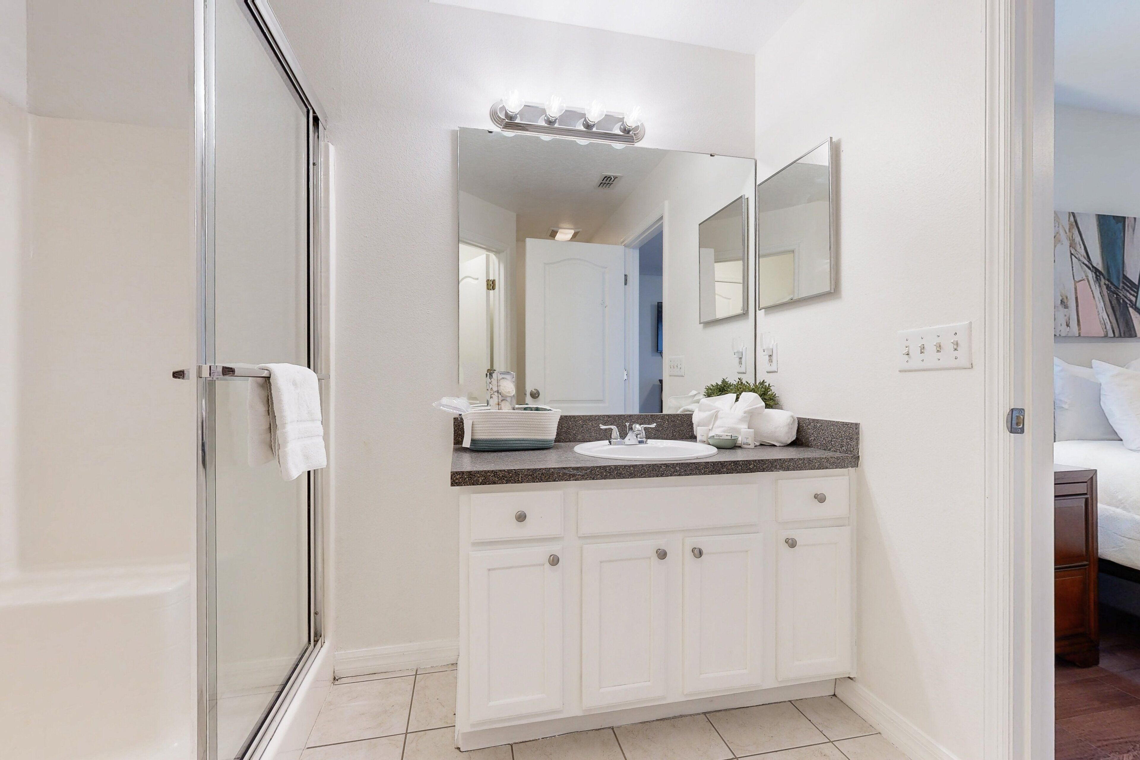 A bathroom with a glass shower door, white cabinets, a sink with a mirror above, and a vanity with a plant.