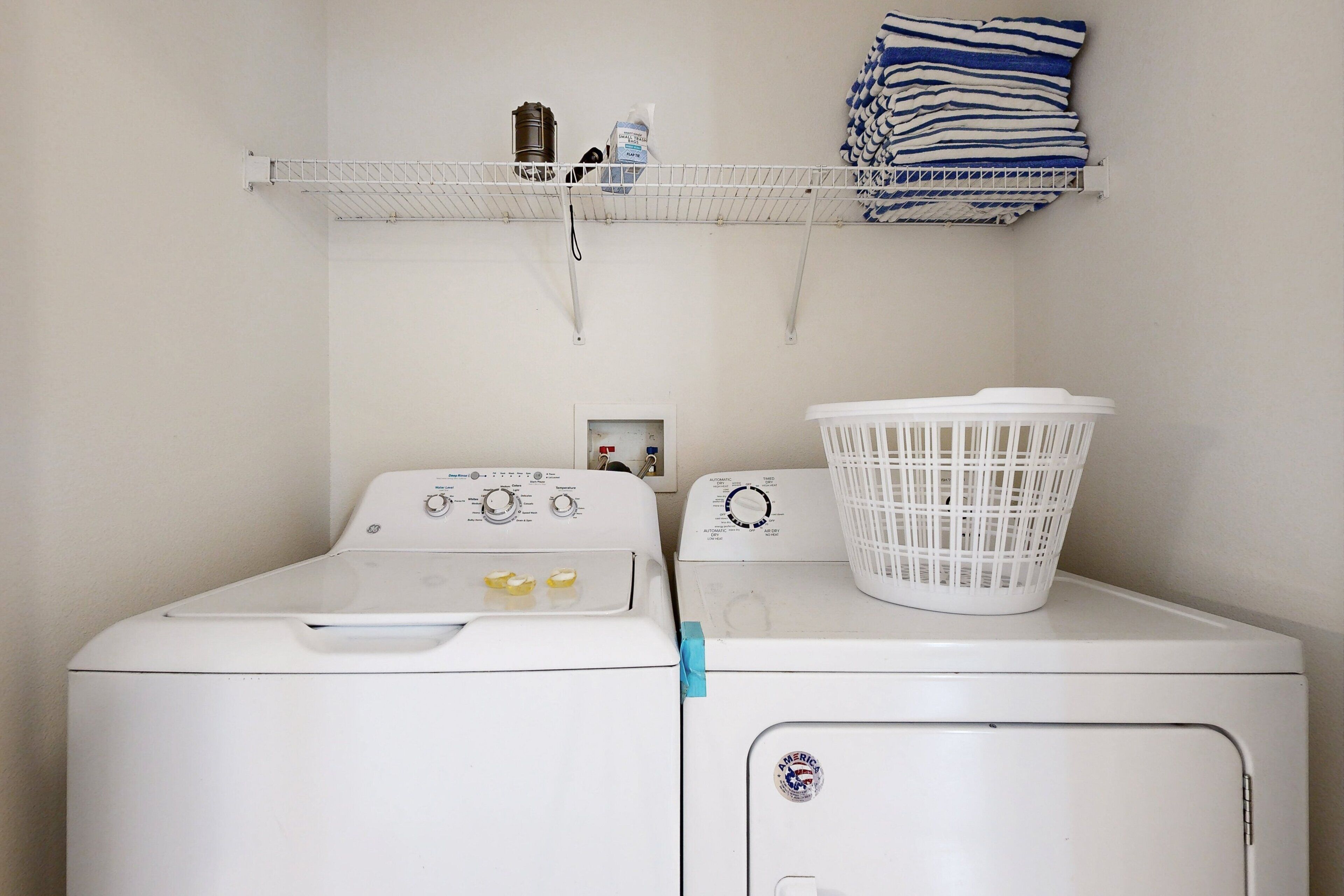 A laundry area with two white washing machines, a white laundry basket, and a shelf with folded towels and a water bottle.