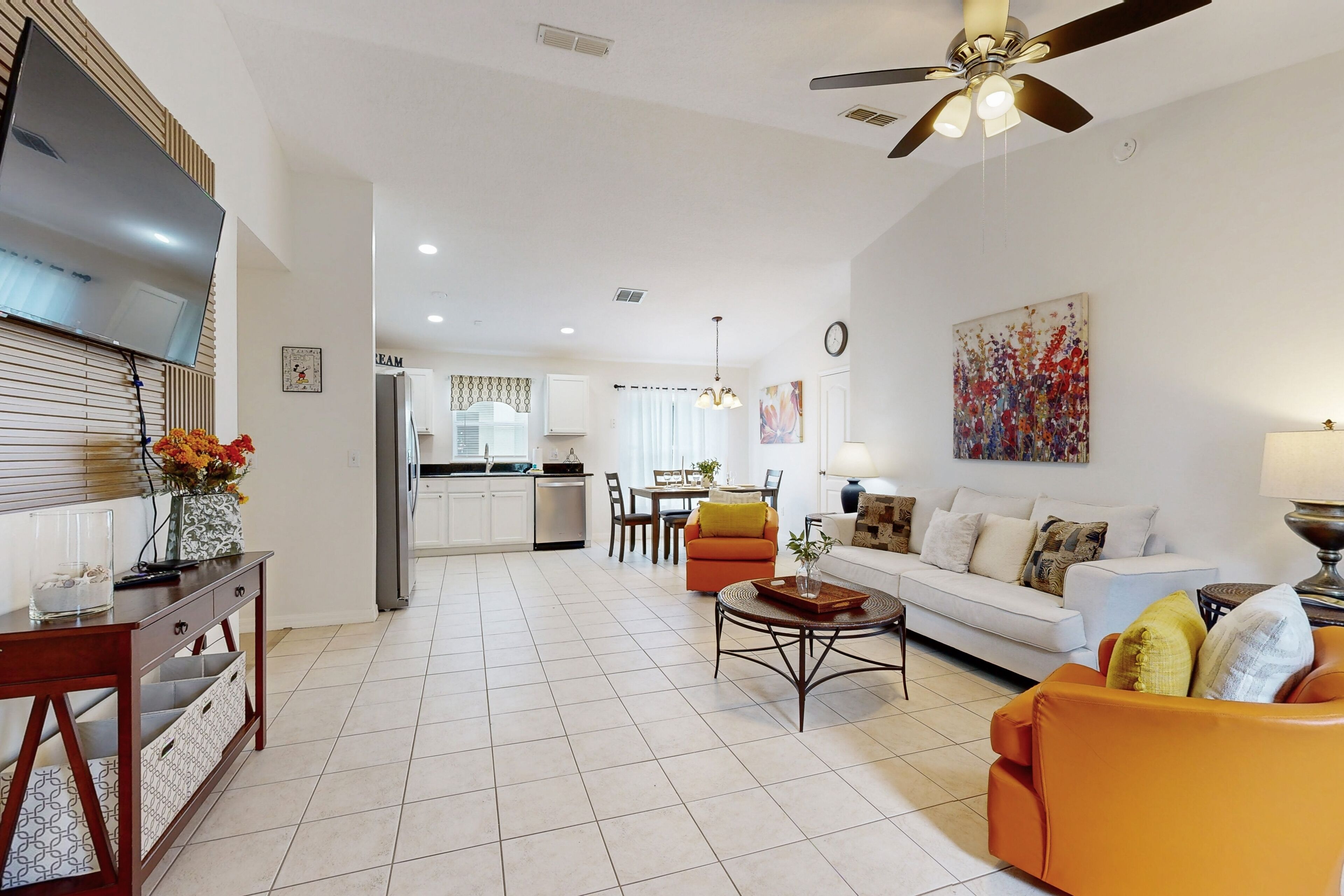 A spacious living room with a sofa, coffee table, and a TV mounted on the wall. There is a kitchen area in the background with a dining table and chairs.