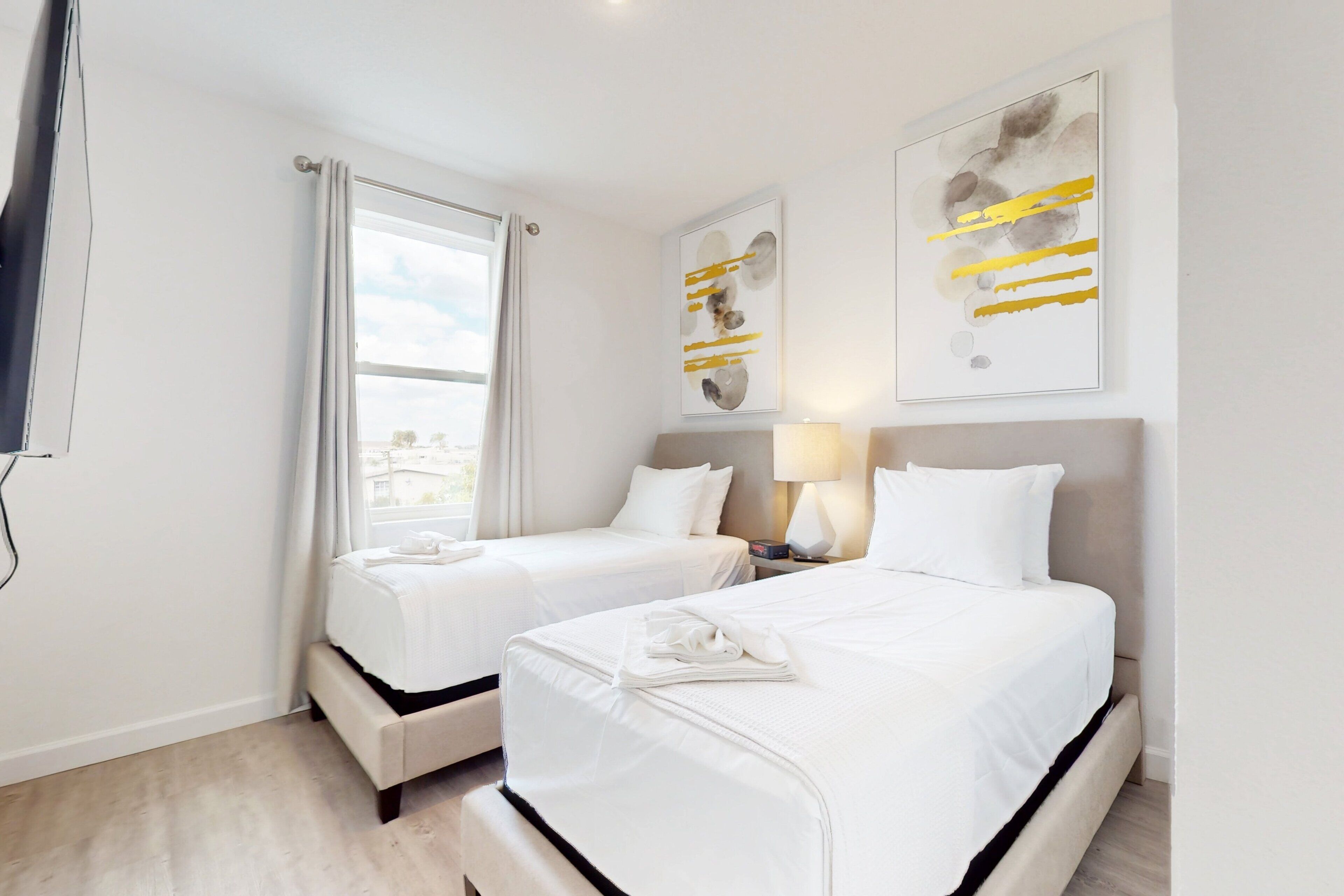 A hotel room with two beds, a window with curtains, and two abstract art pieces on the wall.