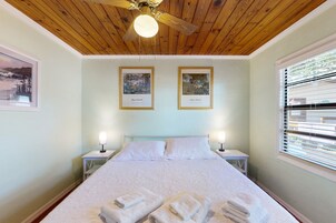 A bedroom with a wooden ceiling, two framed pictures, a bed with white bedding, two bedside lamps, and a window with blinds.