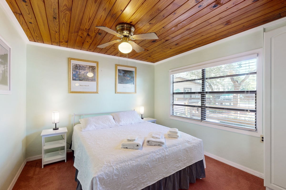 Un dormitorio con techo de madera, una cama con ropa de cama blanca, dos cuadros enmarcados en la pared y una ventana con persianas.
