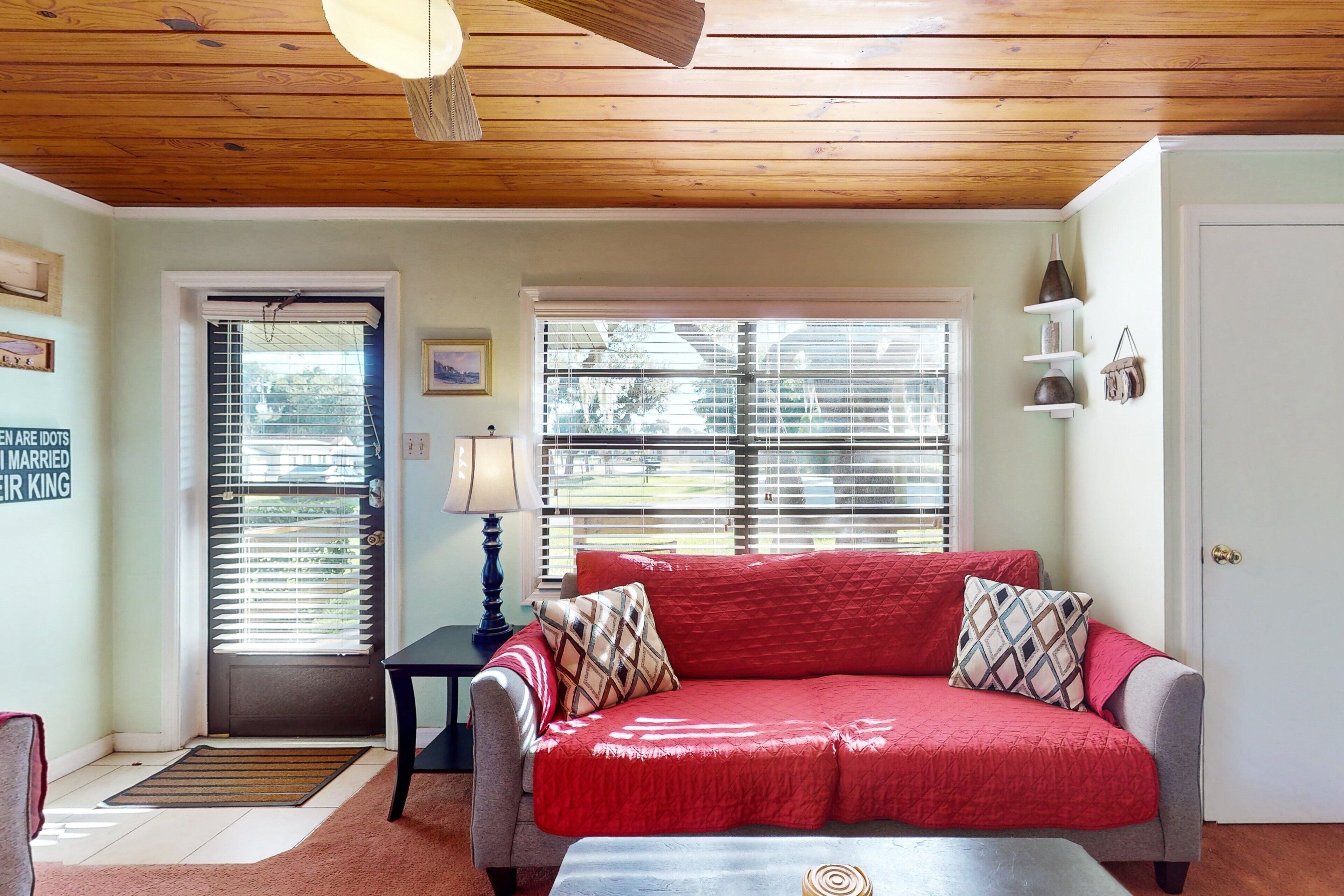A living room with a red sofa, a small table, a lamp, and a door with a doormat.