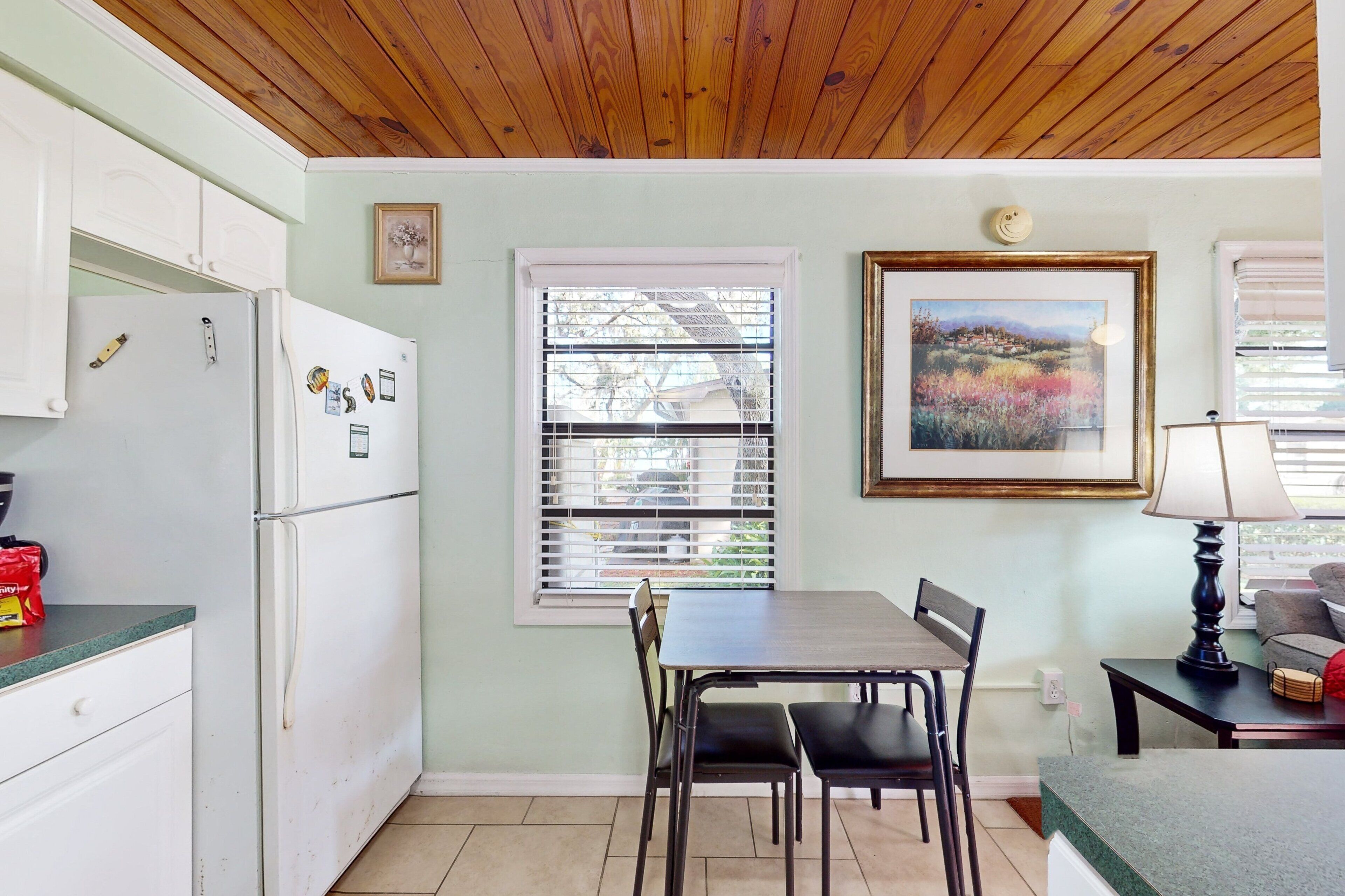 A kitchen with a refrigerator, a dining table with chairs, a window with blinds, and a framed picture on the wall.