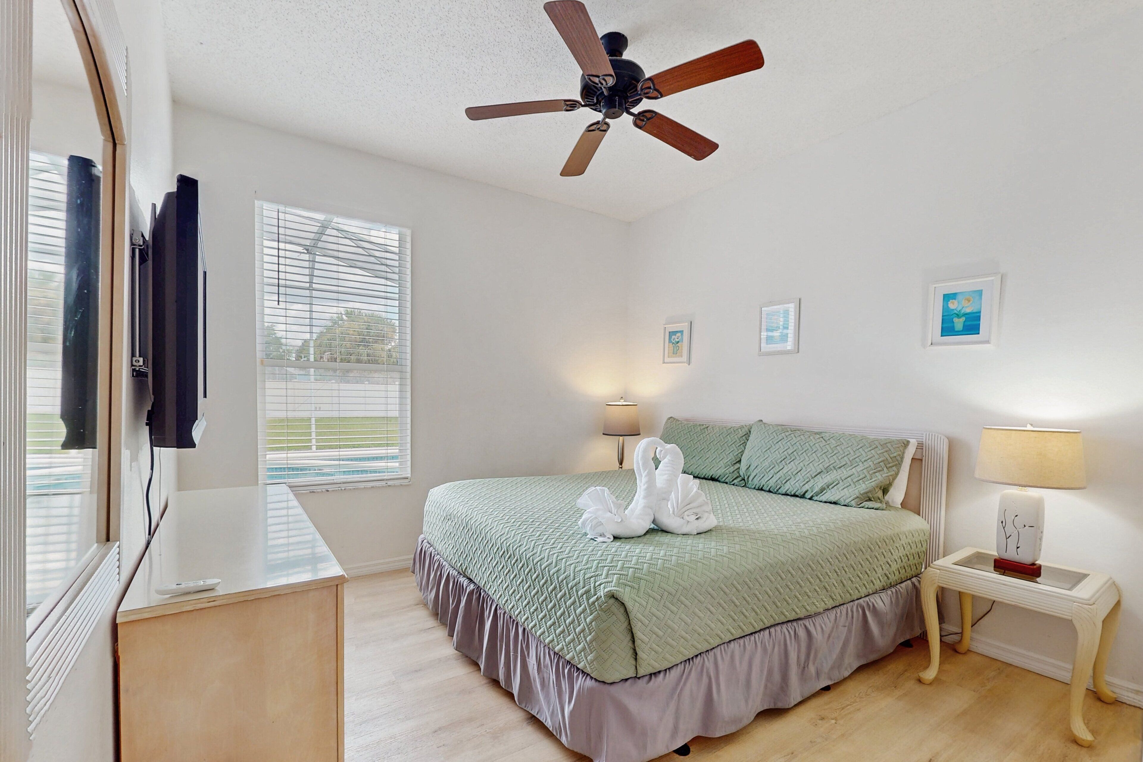 Un dormitorio con cama, ventilador de techo, mesita de noche con lámpara y ventana con persianas.