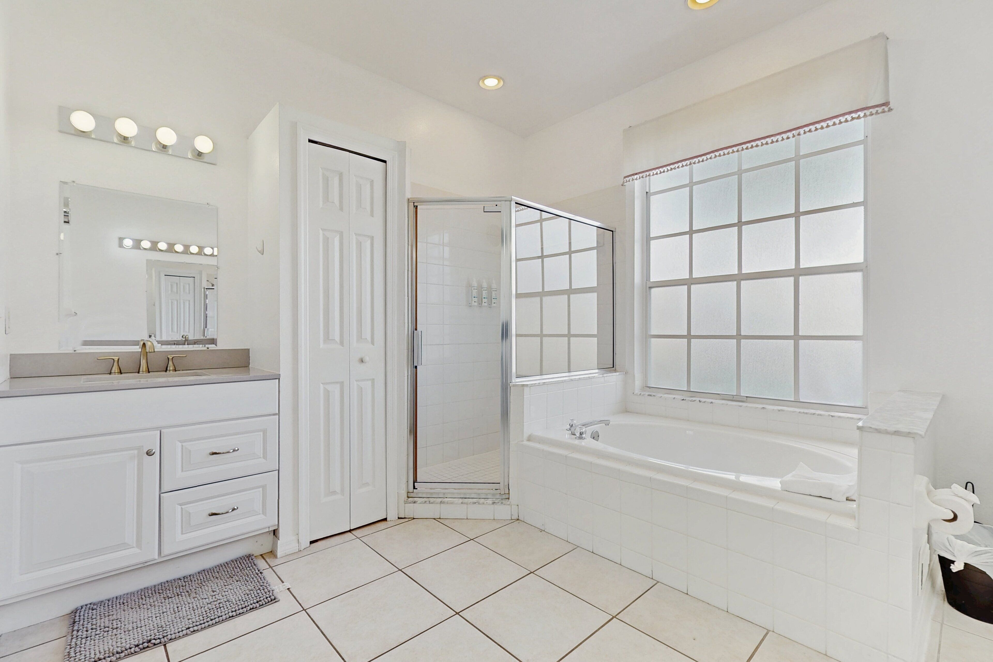 A bathroom with a white vanity, a large mirror with lights, a bathtub, and a shower enclosure.