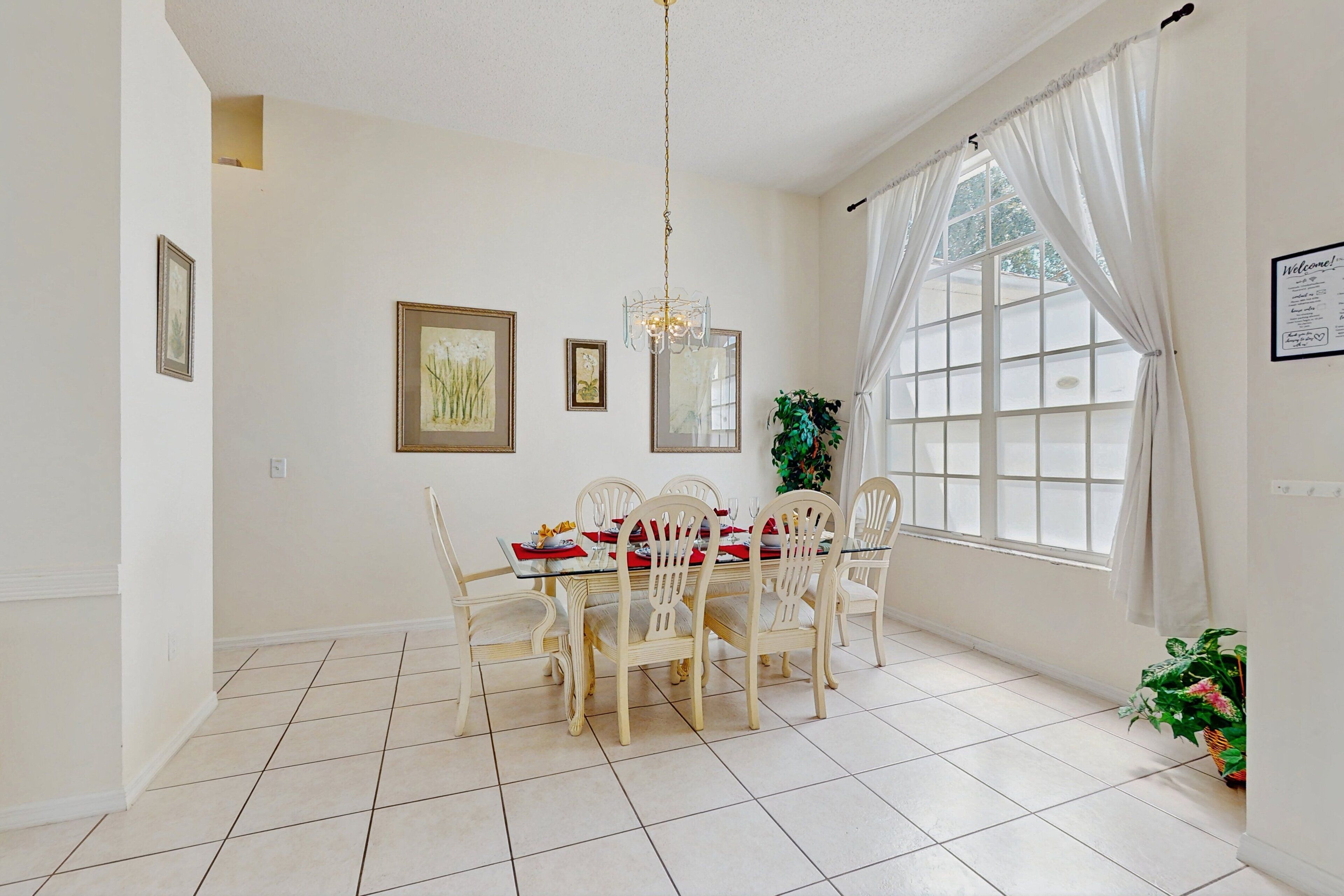 A dining area with a table set for six, white chairs, a chandelier, and a large window with curtains.