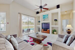 Ein geräumiges Wohnzimmer mit großem Fernseher, Deckenventilator und Blick auf einen Pool.