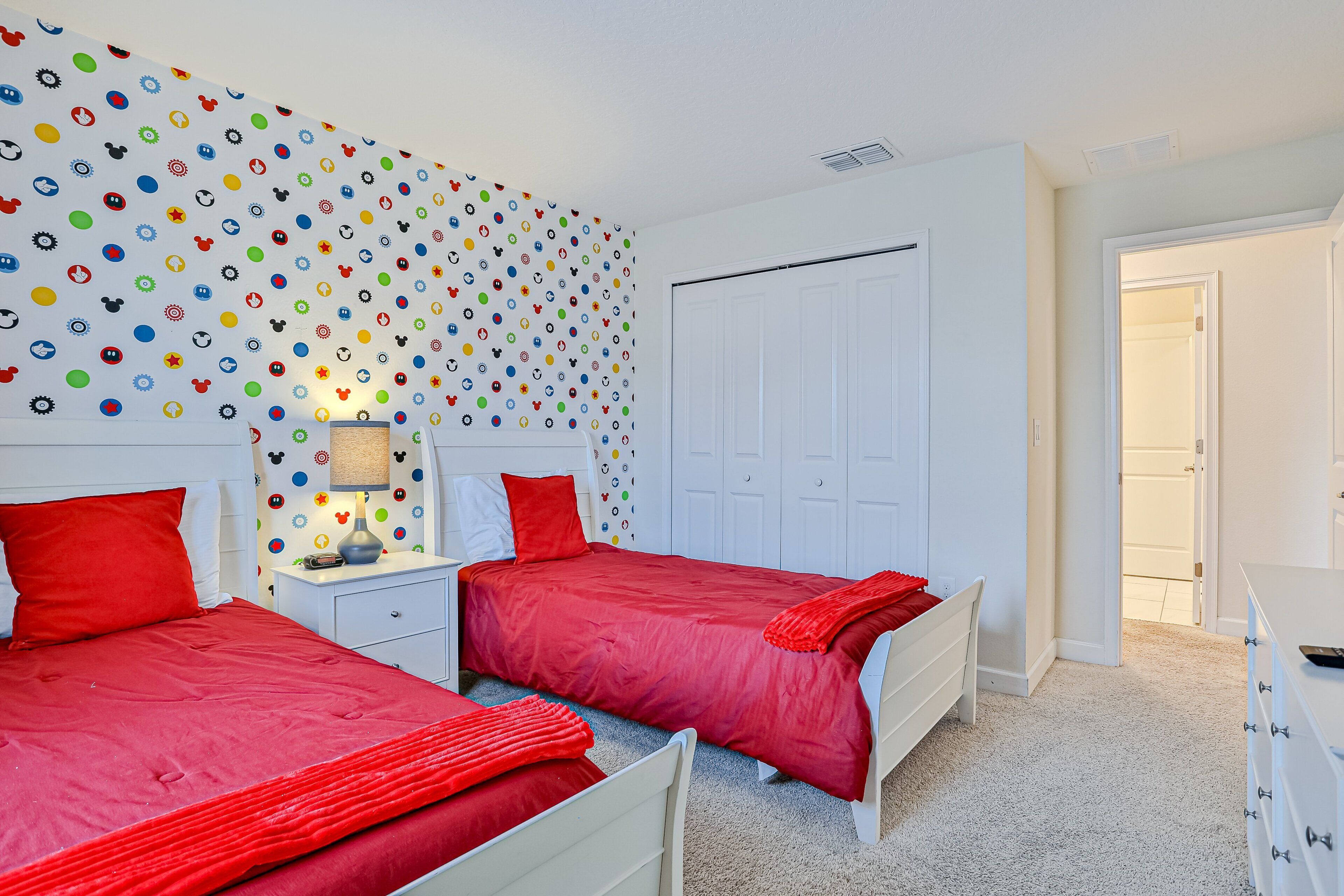 A bedroom with two beds, a white dresser, a nightstand with a lamp, and a wall with colorful circular patterns.