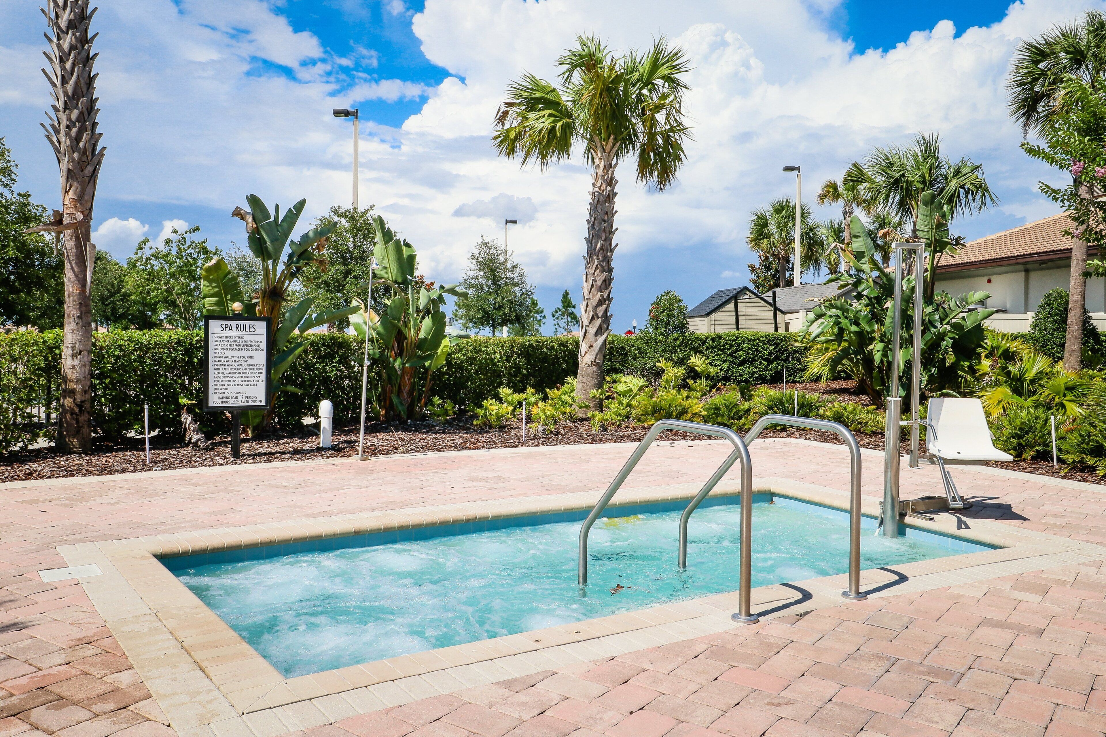 A swimming pool with a sign indicating spa rules, surrounded by palm trees and a brick pathway.