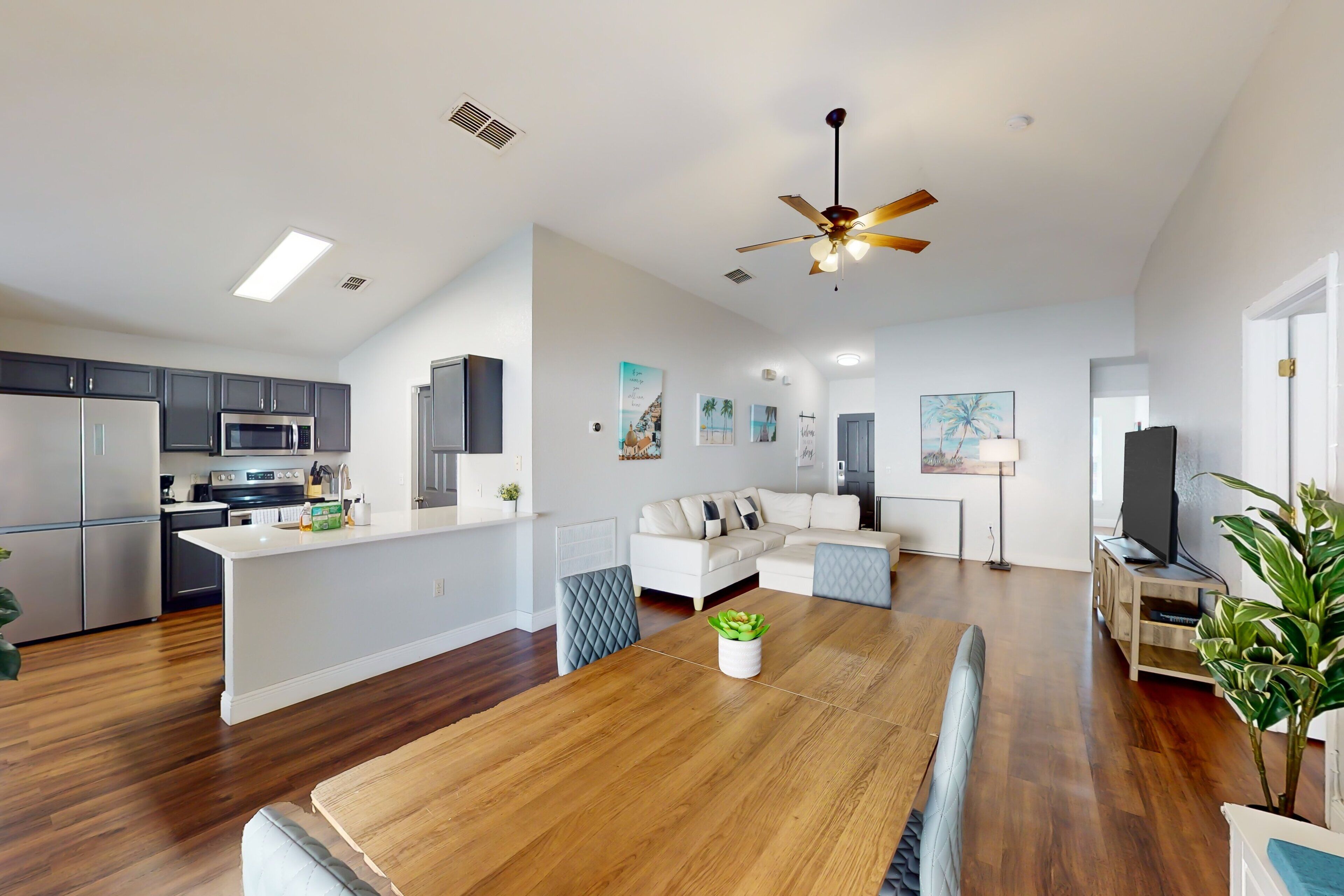 A modern kitchen with stainless steel appliances, a dining area with a wooden table, and a living room with a sofa and a ceiling fan.