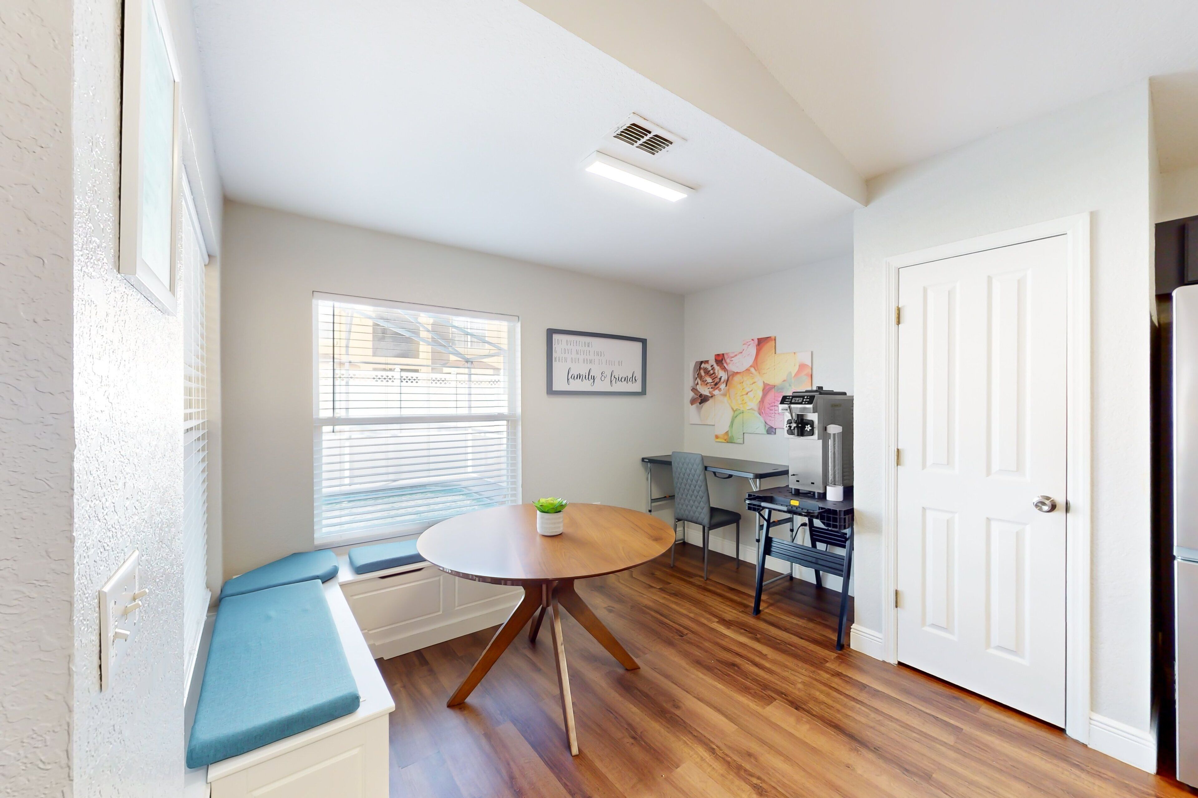 A room with a round wooden table, a bench with cushions, a window with blinds, a door, and a kitchen area with a refrigerator.
