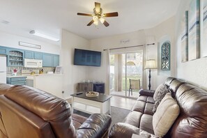 A living room with a brown leather sofa, a coffee table, and a ceiling fan. There is a kitchen area with blue cabinets and a microwave. - Wonderful, Two-level Home Close to Theme Parks With W/d, Central AC & Patio (Davenport)