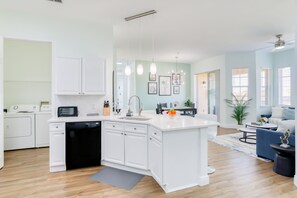 A modern kitchen with white cabinetry, a dishwasher, and a built-in oven. There is a dining area with a blue sofa and a small table. - Snowbird-friendly Condo Near Walt Disney World Resort W/pool, Wifi, AC, and W/D (Davenport)
