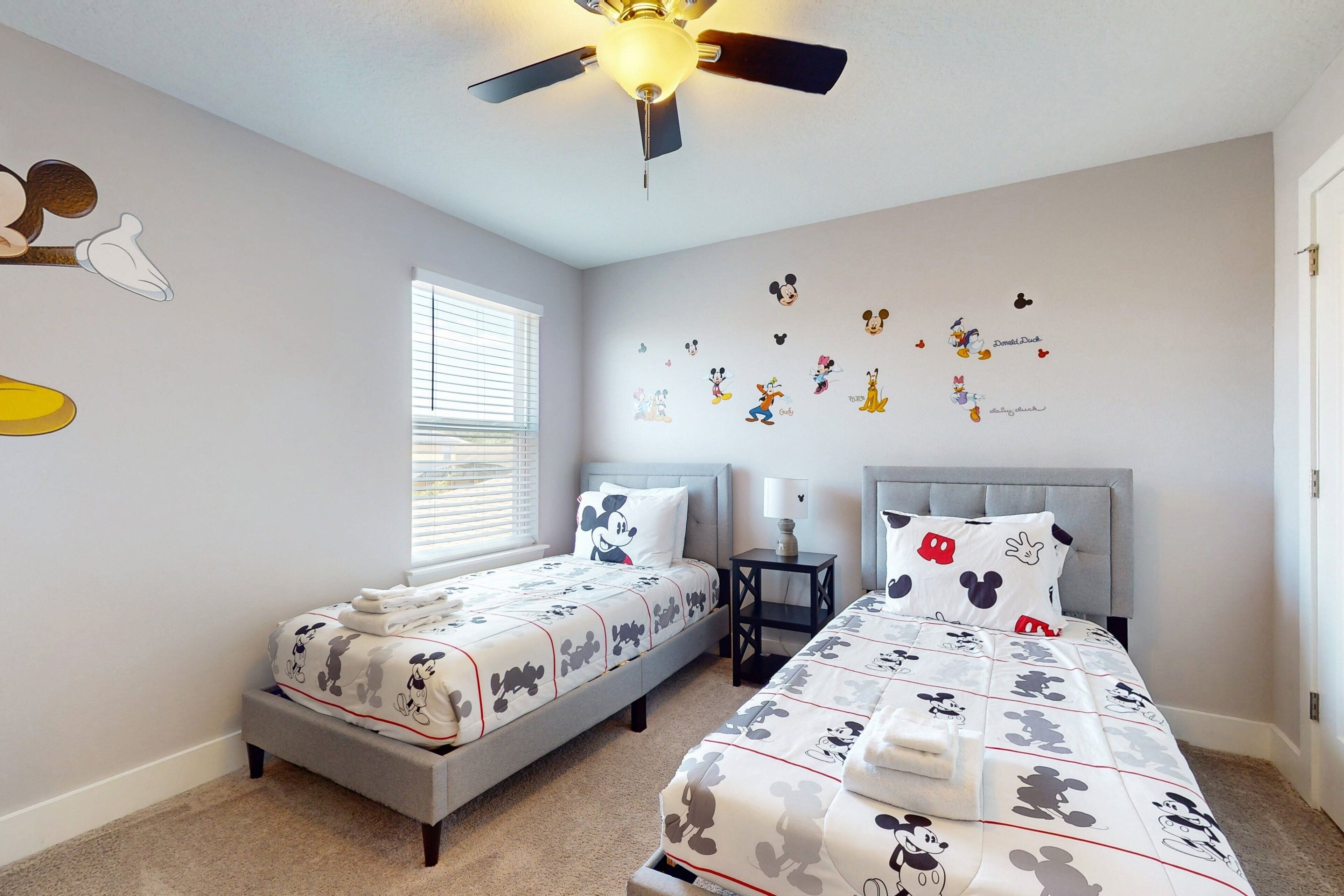 Une chambre avec deux lits, un ventilateur de plafond et des autocollants de Mickey Mouse sur le mur.