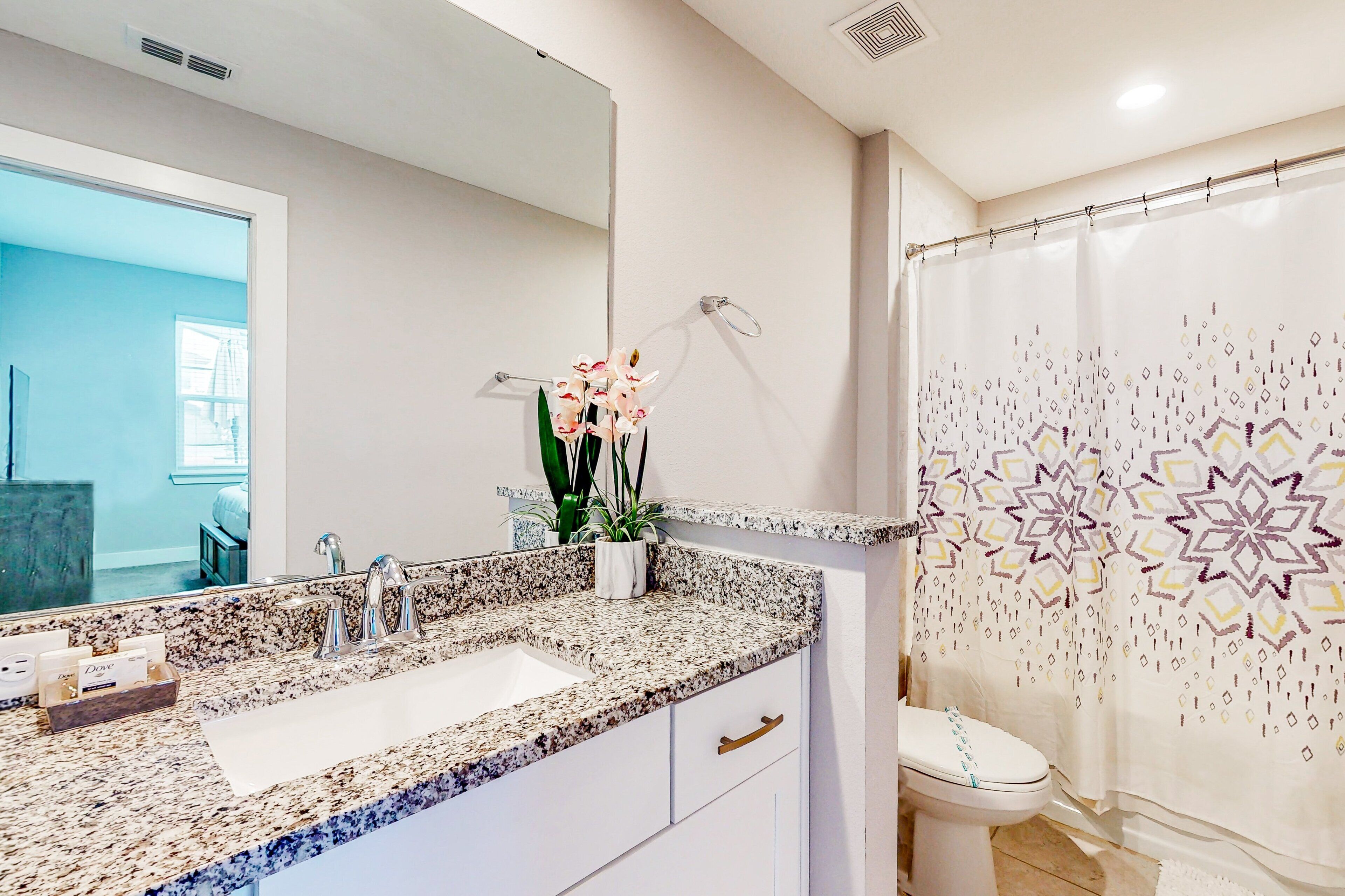 A bathroom with a granite countertop, a sink, a toilet, a shower curtain with a geometric pattern, and a potted orchid.