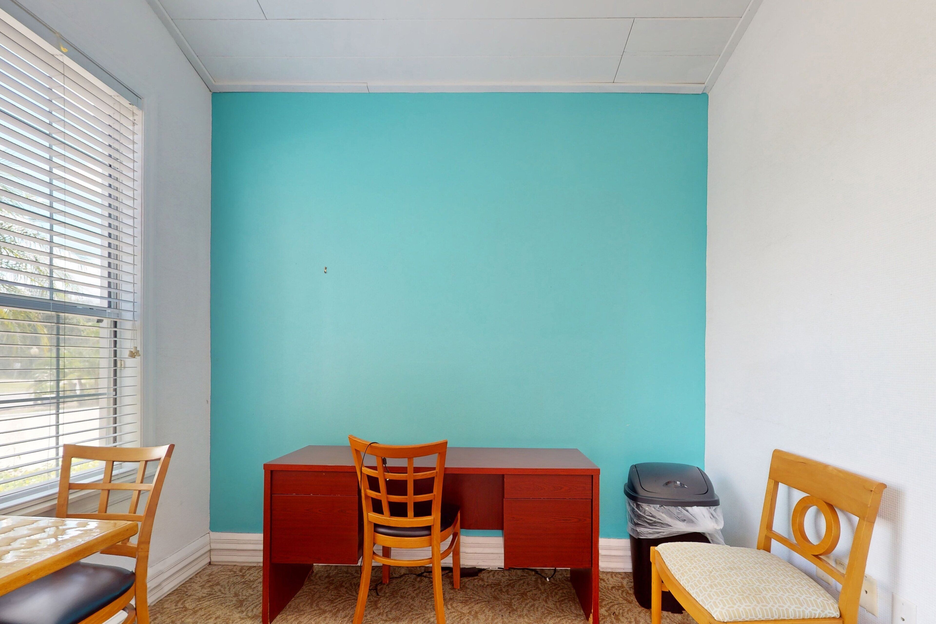 A room with a teal wall, a wooden desk, two chairs, and a trash bin.