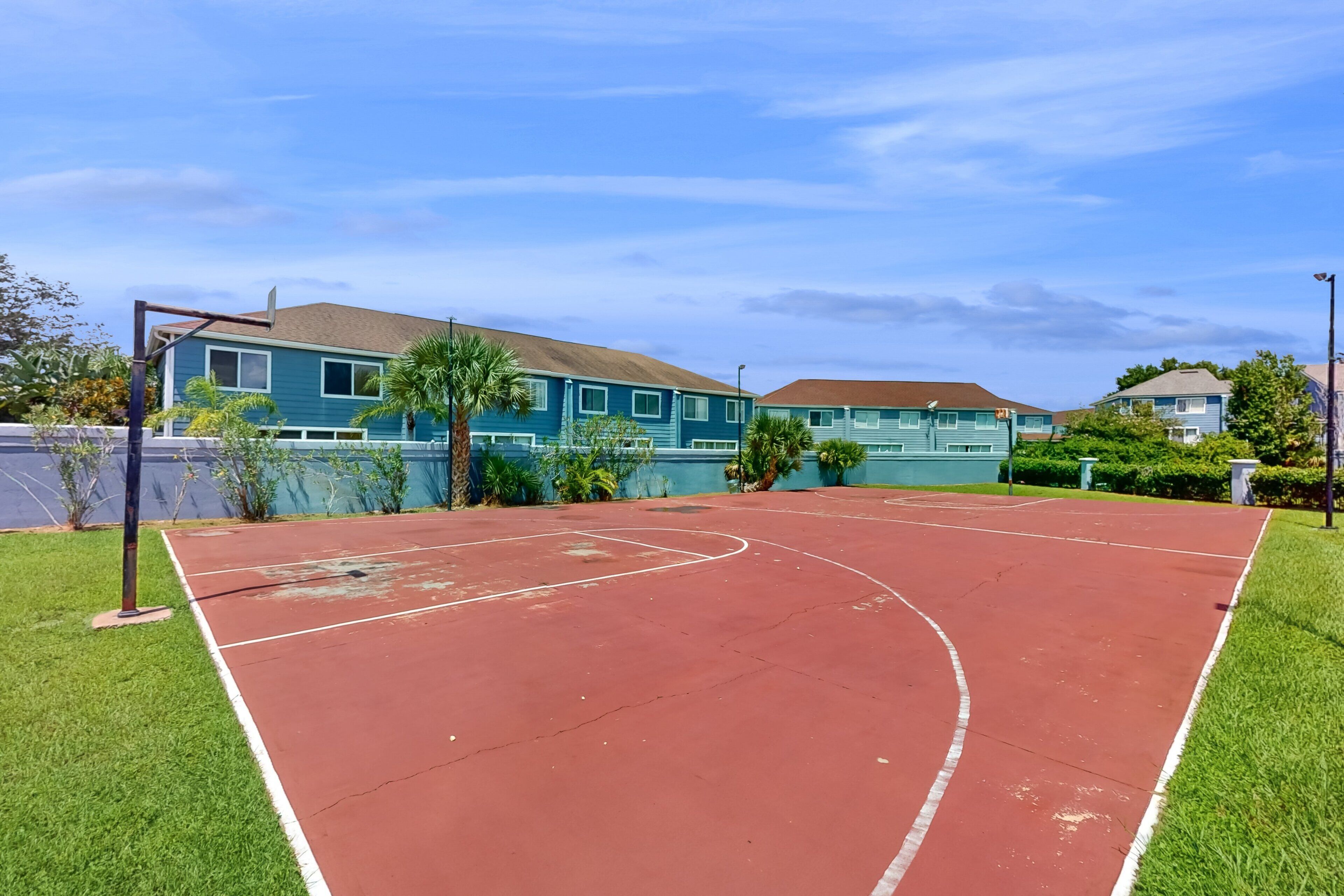 Una cancha de baloncesto al aire libre con superficie roja, rodeada de césped verde y palmeras, con casas al fondo.