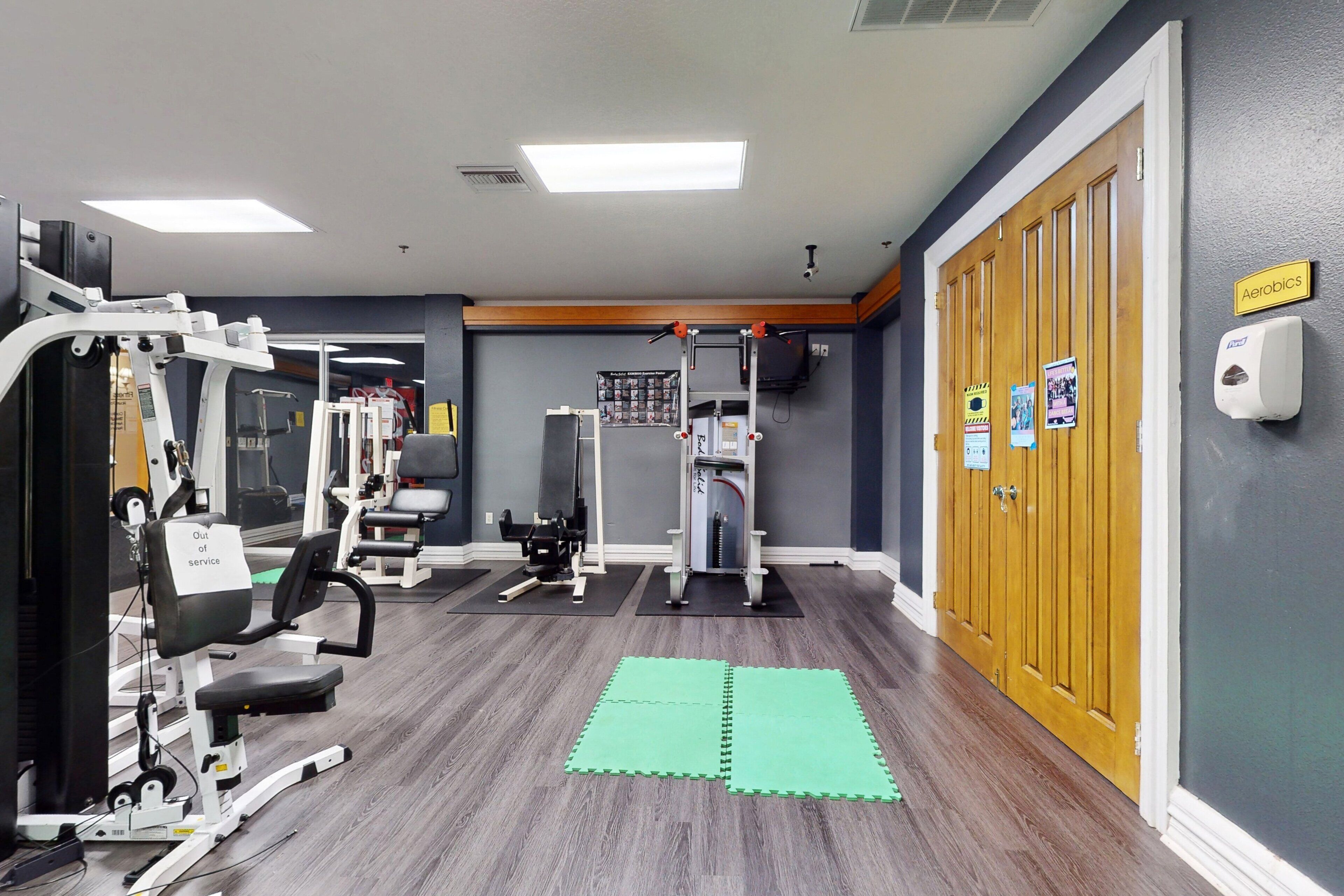 Un gimnasio con equipo de ejercicio, colchonetas verdes y una puerta de madera con la leyenda 'Aerobics'.