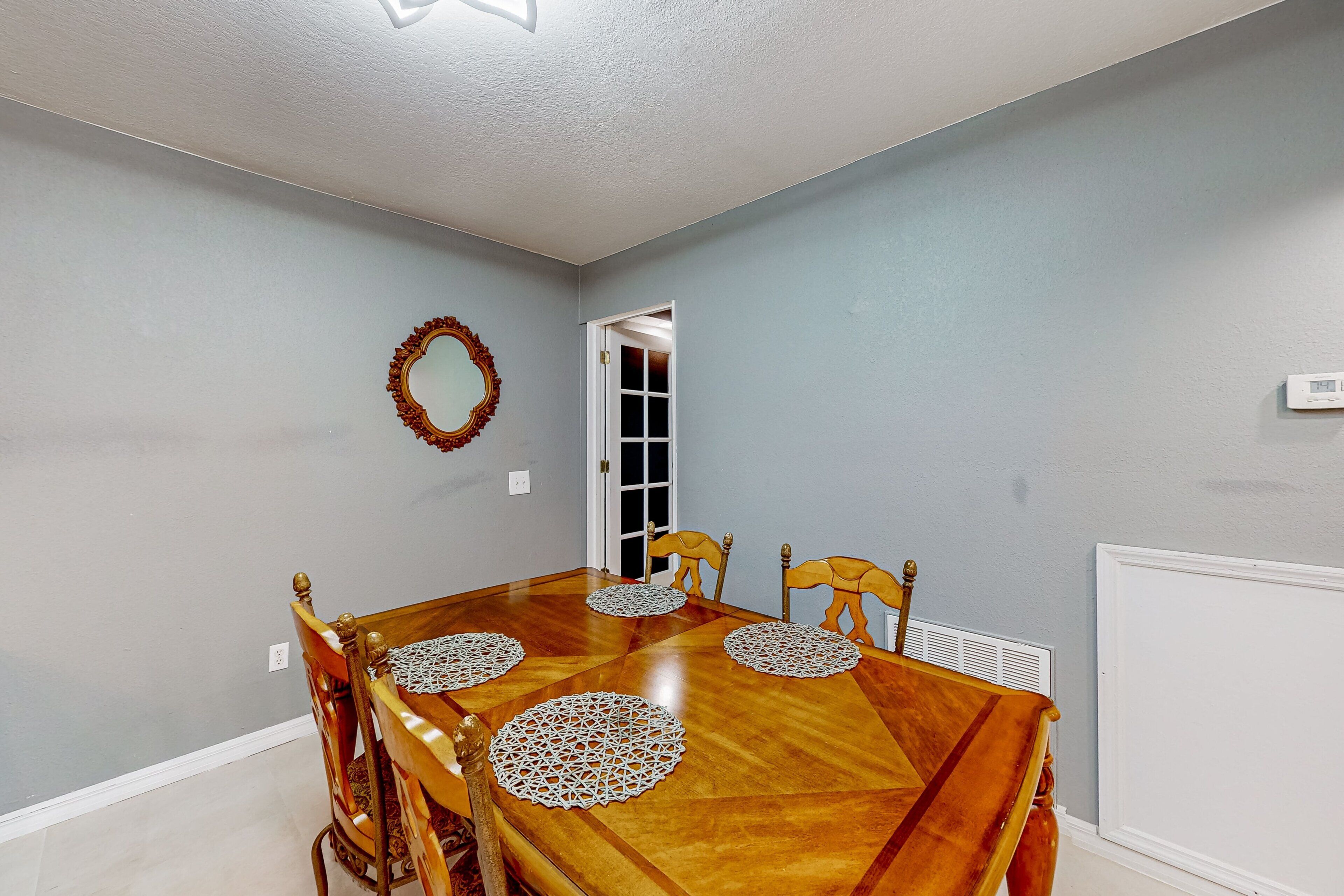 Une salle à manger avec une table en bois, quatre chaises, un miroir décoratif et une porte donnant sur une autre pièce.
