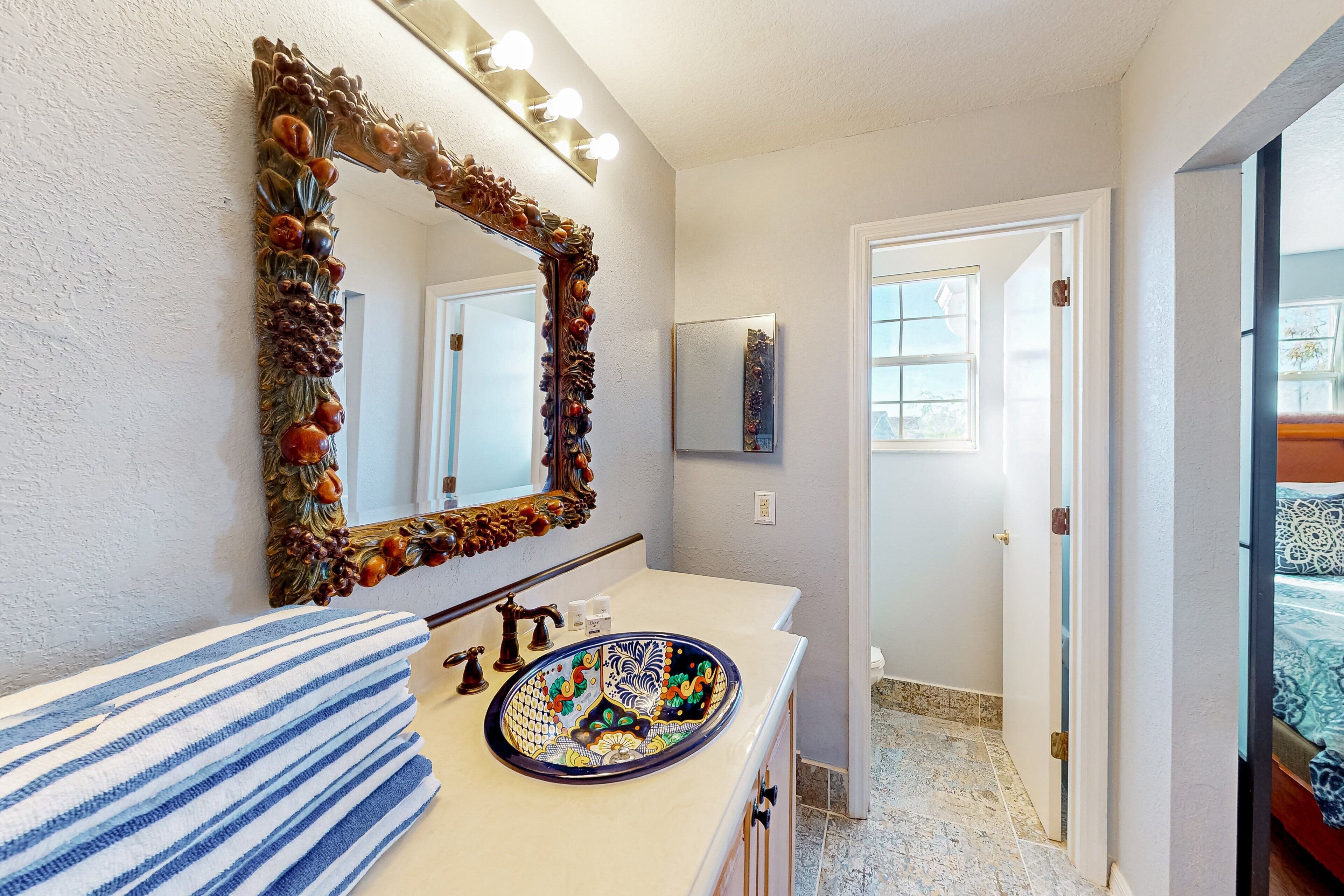 Une salle de bain avec un miroir décoratif, un lavabo orné d’un récipient aux couleurs vives et une serviette à rayures.