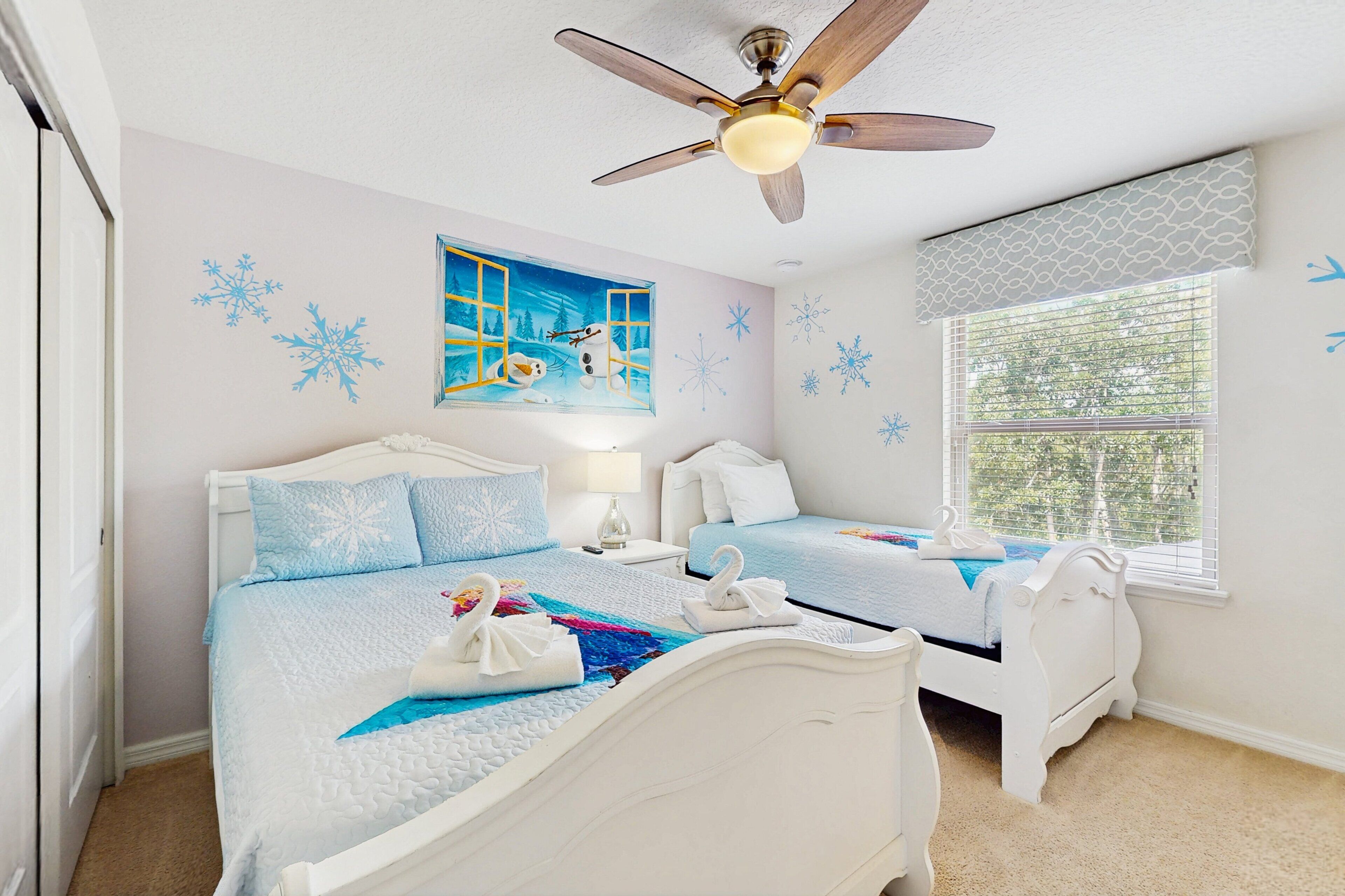A bedroom with two beds, a ceiling fan, a window with blinds, and a wall with snowflake decorations.