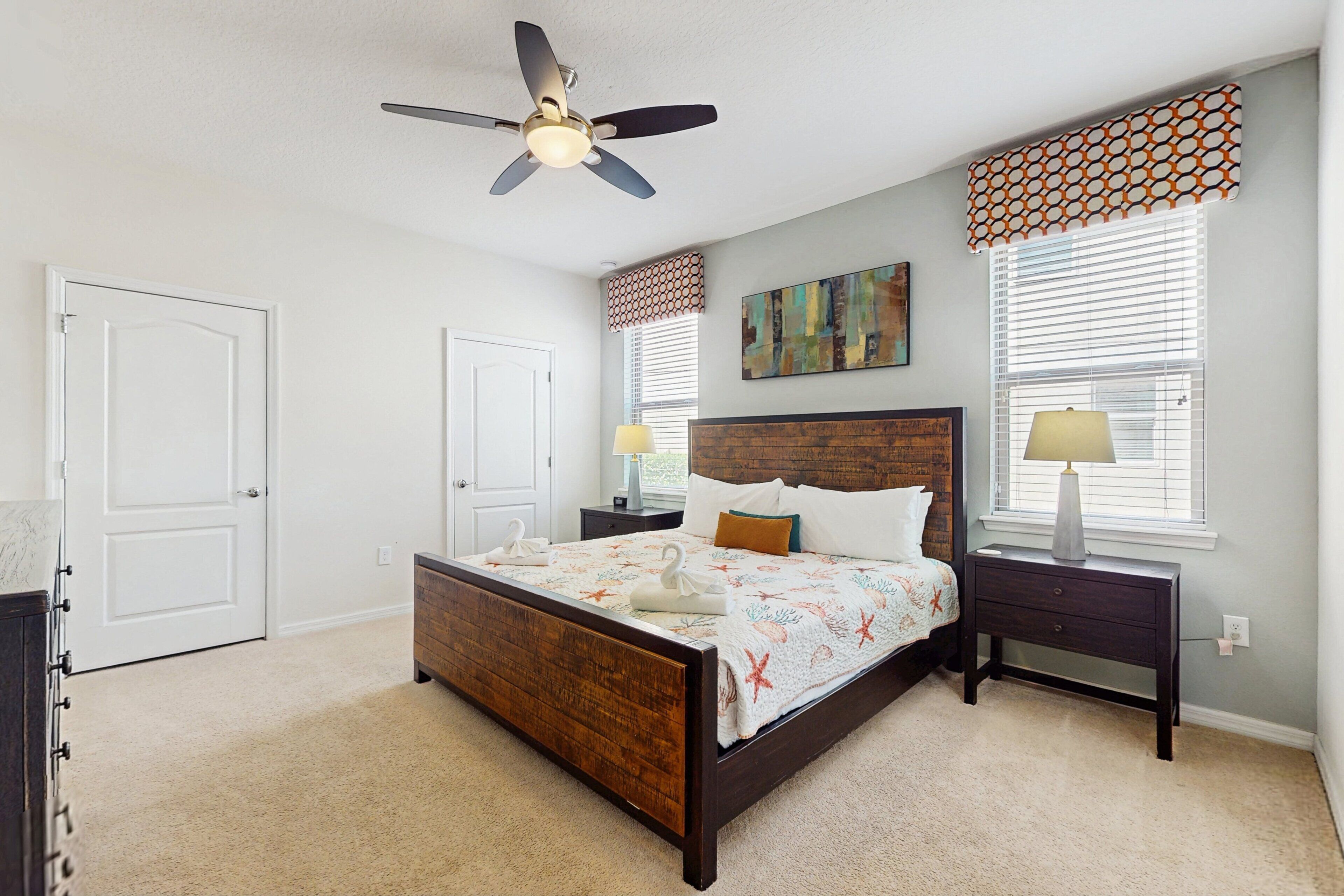 A bedroom with a wooden bed, two bedside tables with lamps, a ceiling fan, and a large window with blinds.