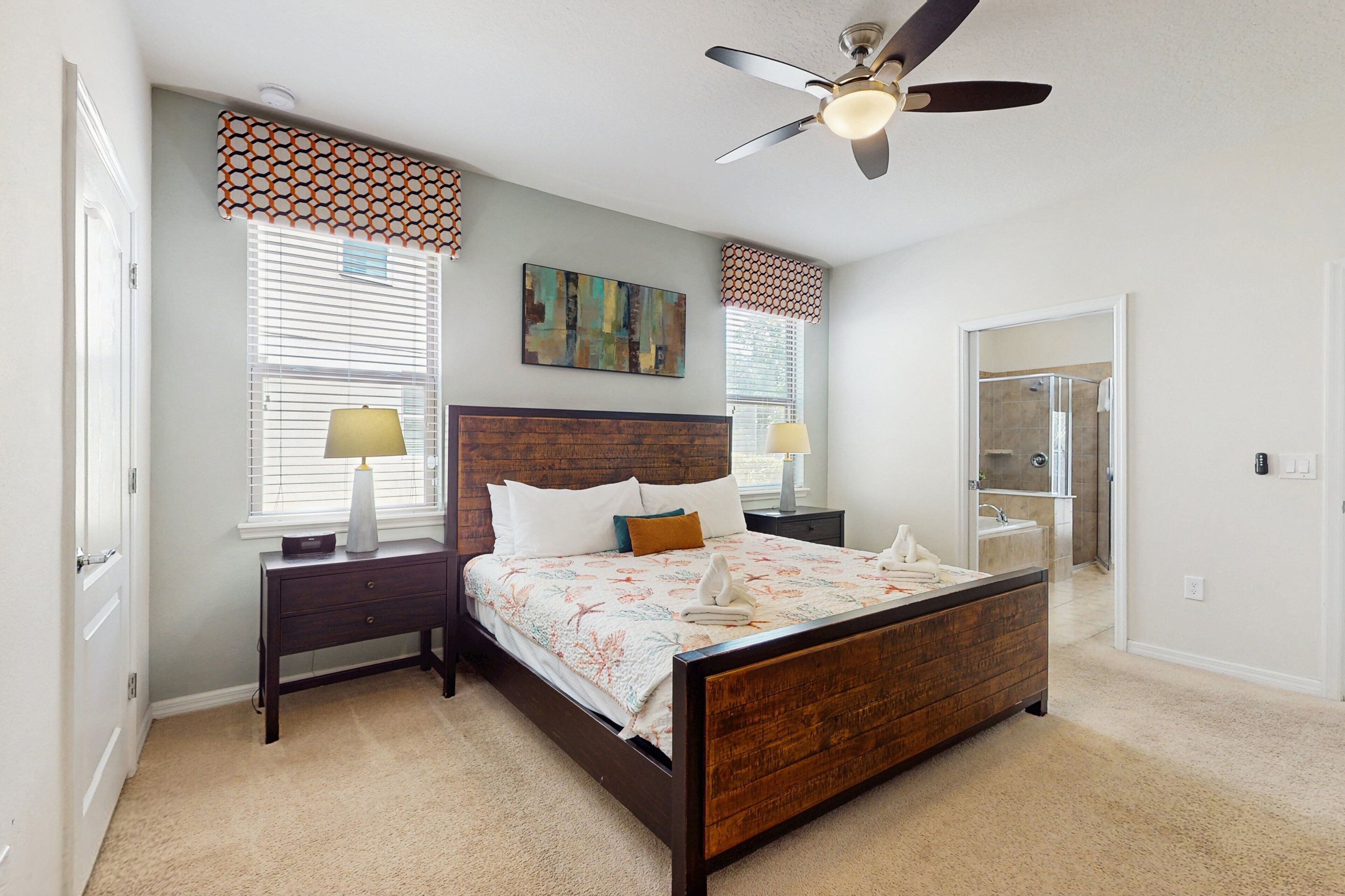 A bedroom with a wooden bed, a ceiling fan, a window with blinds, a nightstand, and a bathroom visible through an open door.