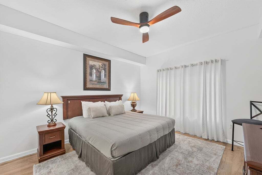 A bedroom with a bed, two bedside tables with lamps, a ceiling fan, a painting on the wall, and a window with curtains.