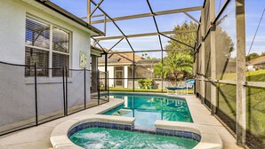 A covered outdoor swimming pool with a hot tub, surrounded by a screened area and a house in the background. - Vibrant Modern Home With Private Pool, Spa, Lanai & Expansive Game Room (Haines City)