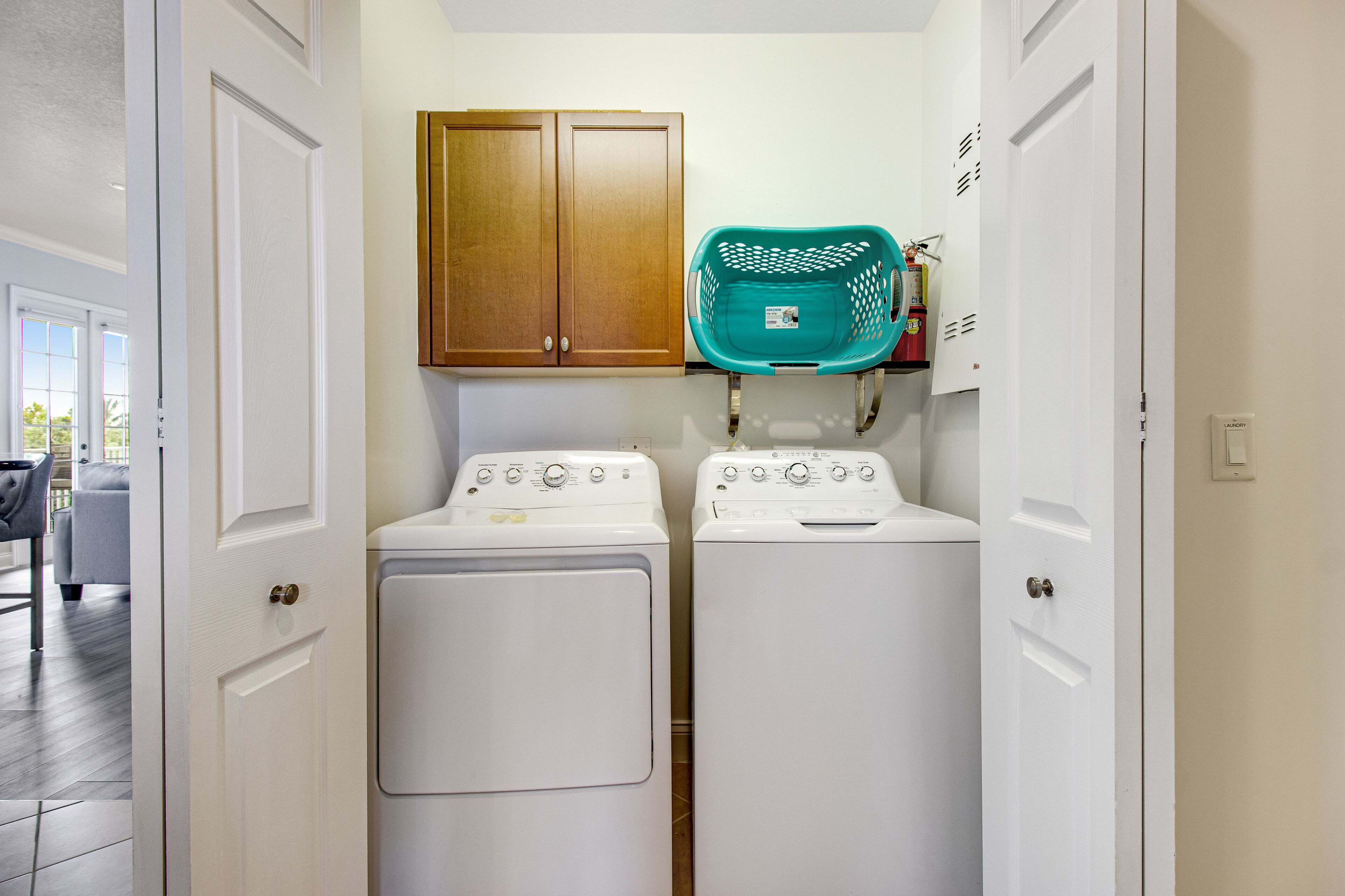 A laundry room with two white washing machines, a wooden cabinet, and a teal laundry basket.