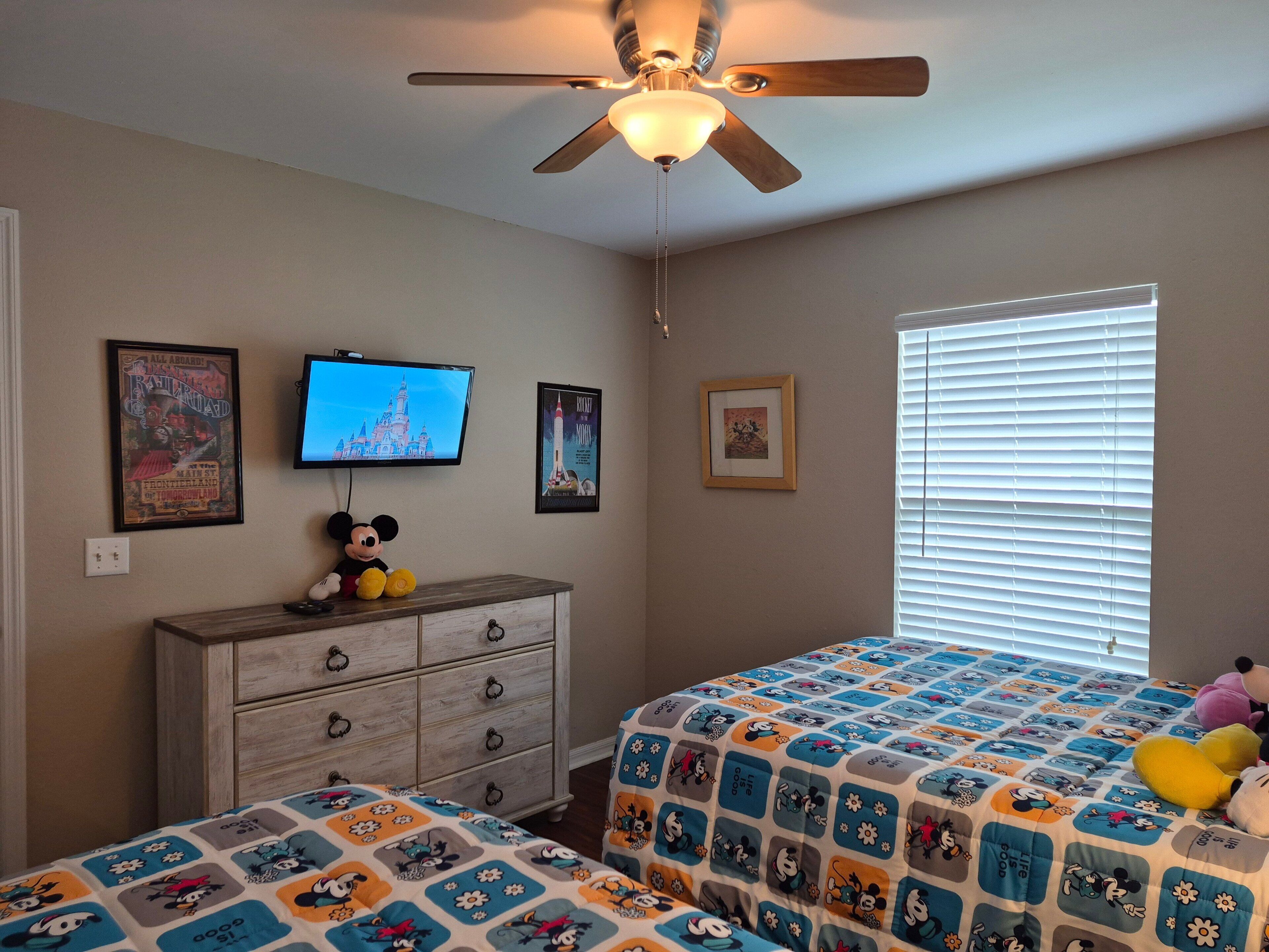 A bedroom with a ceiling fan, a television, and two beds with Mickey Mouse themed bedding.