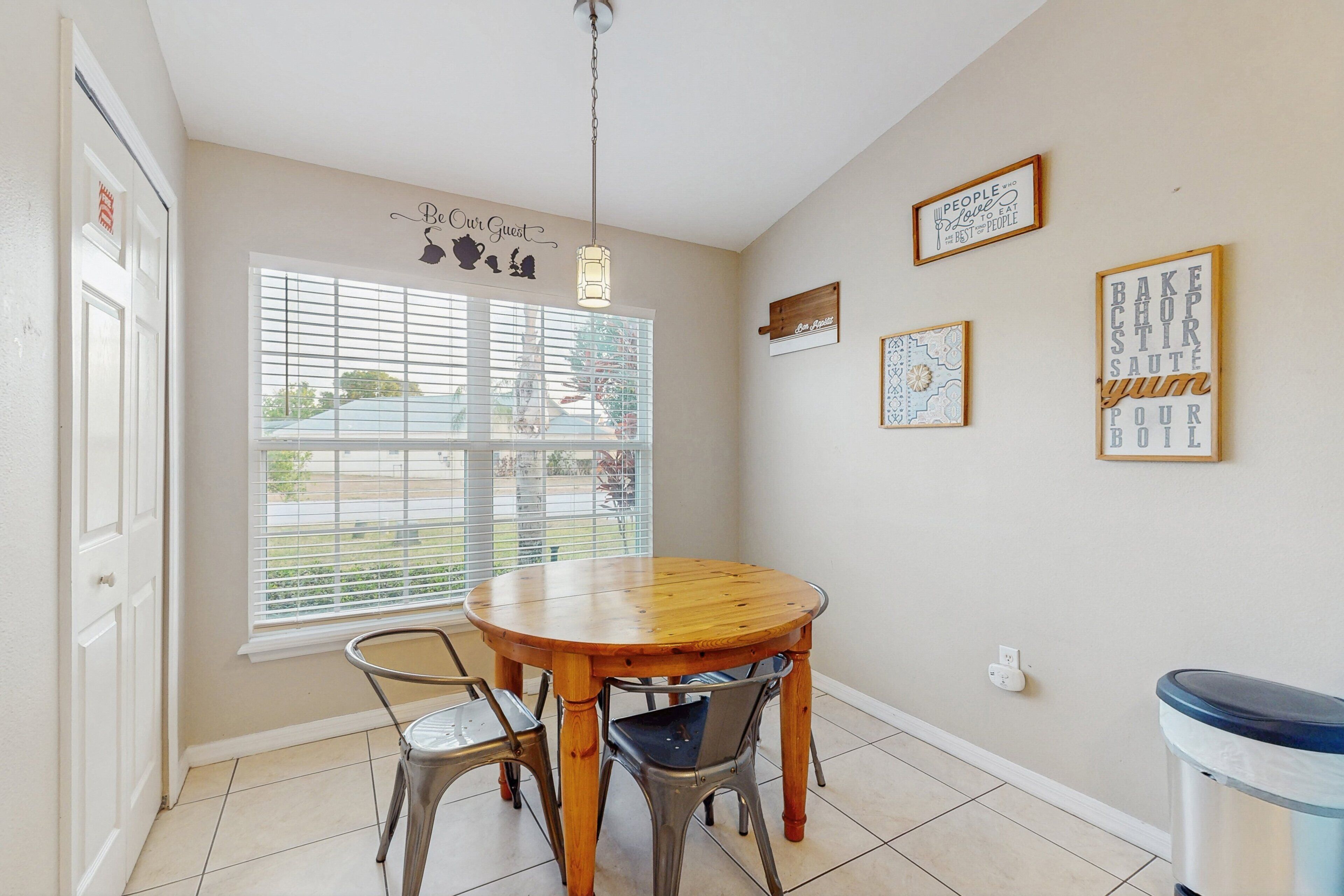 A dining area with a round wooden table, two metal chairs, a window with blinds, and wall decorations including framed text and patterns.
