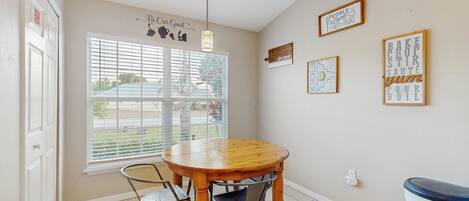 A dining area with a round wooden table, two metal chairs, a window with blinds, and wall decorations including framed text and patterns.