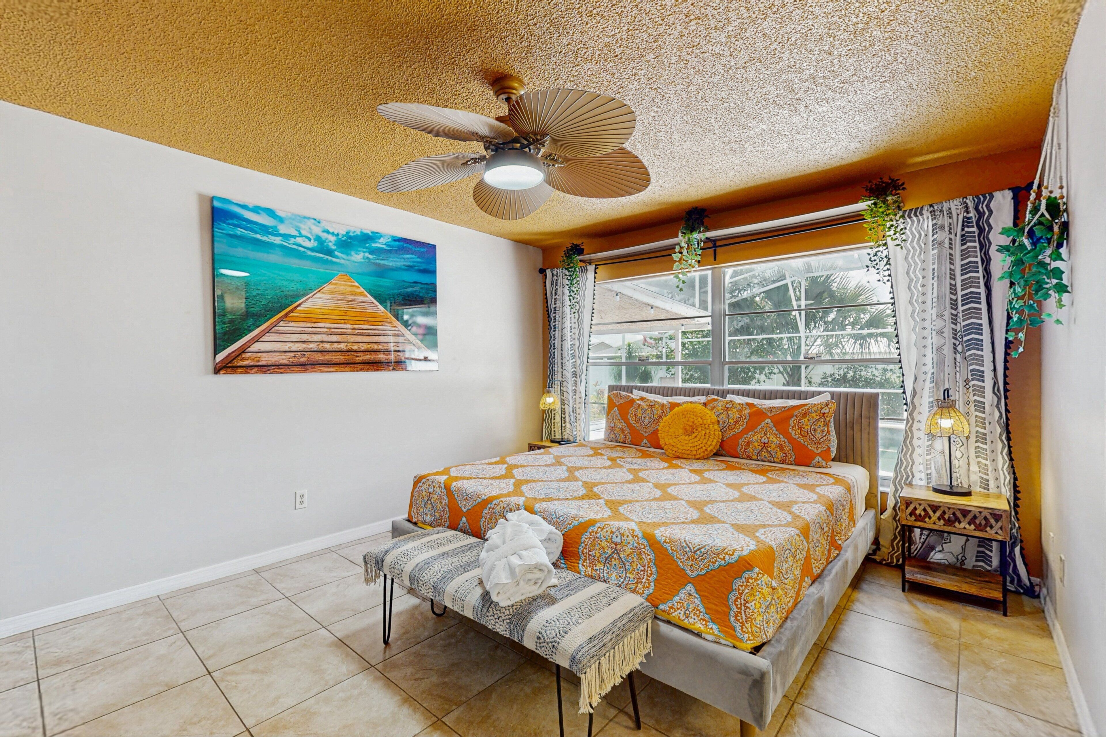 A bedroom with a bed, a bench, a ceiling fan, a window with curtains, and a painting on the wall.